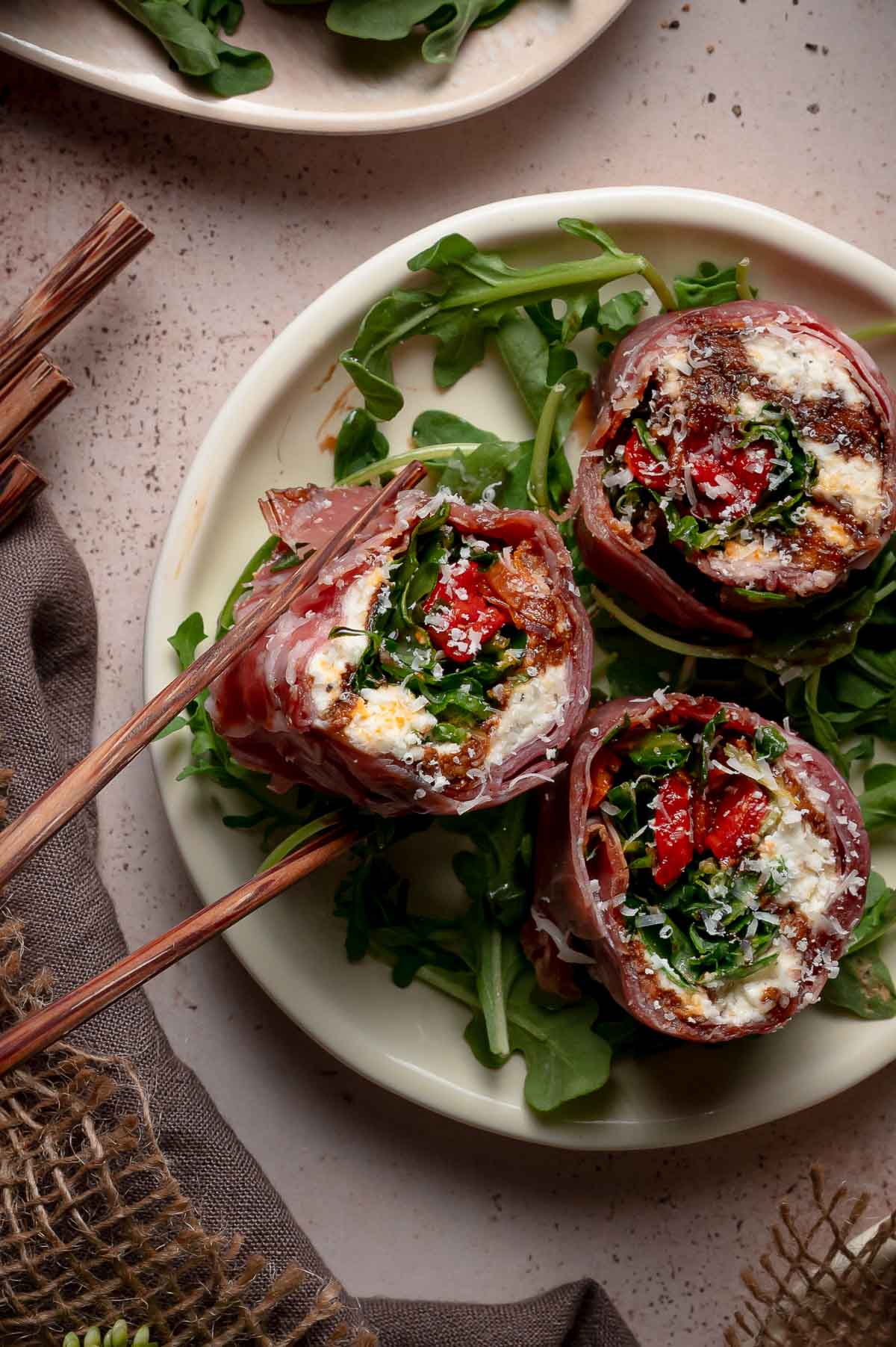 Three prosciutto-wrapped Italian sushi rolls filled with greens, cheese, and roasted red peppers are served on a bed of arugula on a white plate.