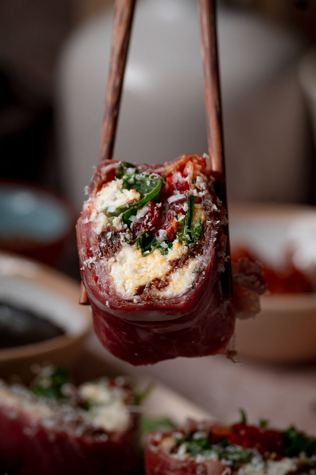 A close-up of chopsticks holding a piece of Italian sushi, filled with cheese wrapped in cured meat.