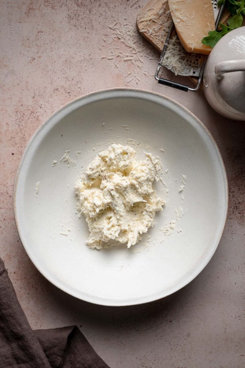 A white plate with a small mound of mascarpone cheese mixed with other ingredients, with a wedge of grated Parmesan and a cheese grater nearby on a light-colored surface.