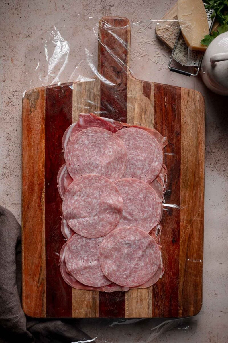 Slices of salami arranged in a rectangle on over other meats and plastic wrap.