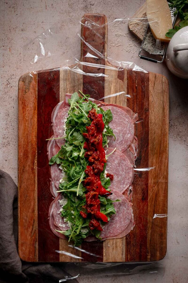A wooden board with overlapping salami, arugula, and chopped sun-dried tomatoes layered over plastic wrap, ready to be rolled.