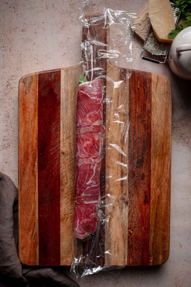 An Italian sushi rolled in plastic wrap is placed on a wooden cutting board.