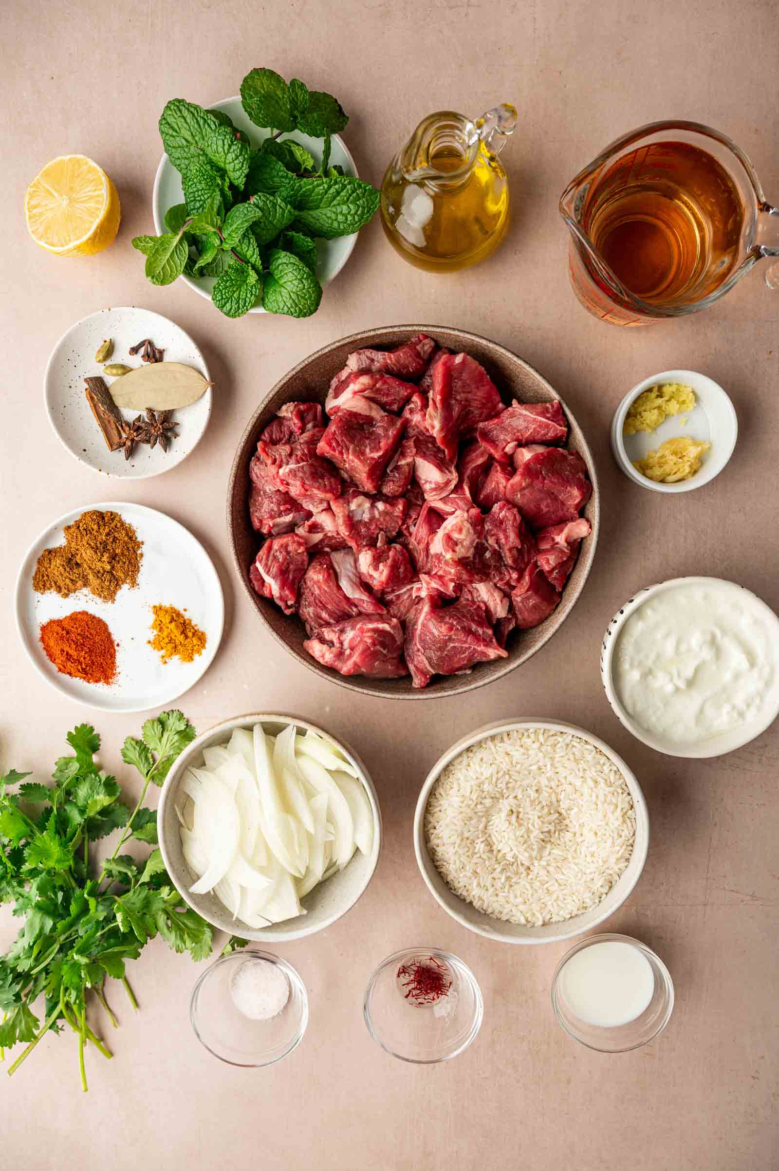 A flat lay of ingredients for a lamb biryani, including raw lamb pieces, rice, sliced onions, yogurt, herbs, spices, oil, ginger, garlic, lemon, and broth on a beige surface.