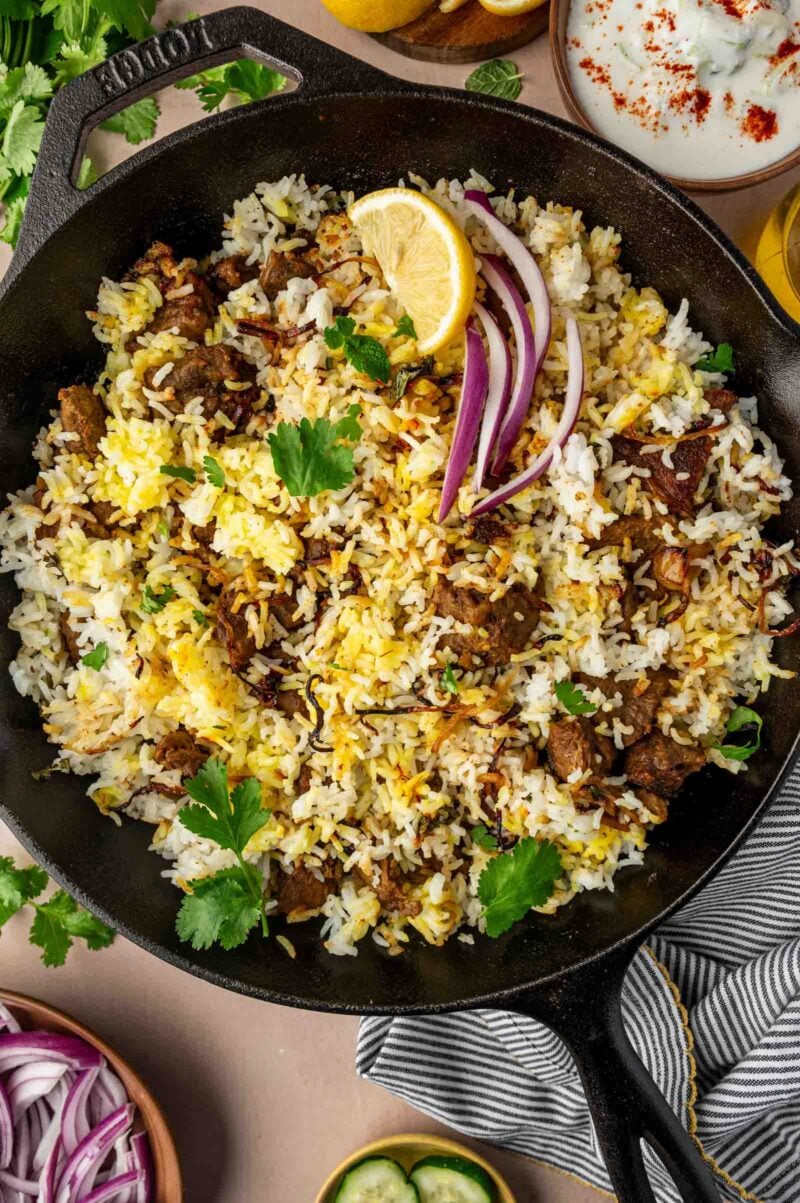 A skillet filled with lamb biryani rice, garnished with cilantro, red onion slices, and a lemon wedge.