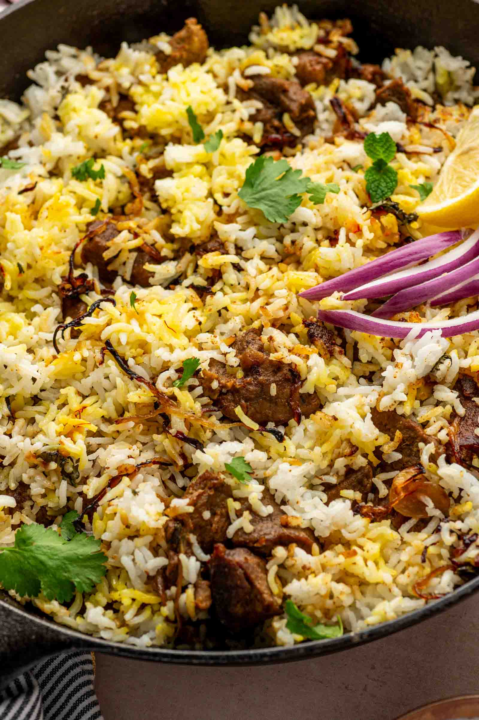 A close-up of biryani with rice and l of cooked lamb, garnished with cilantro, sliced red onions, and a lemon wedge.