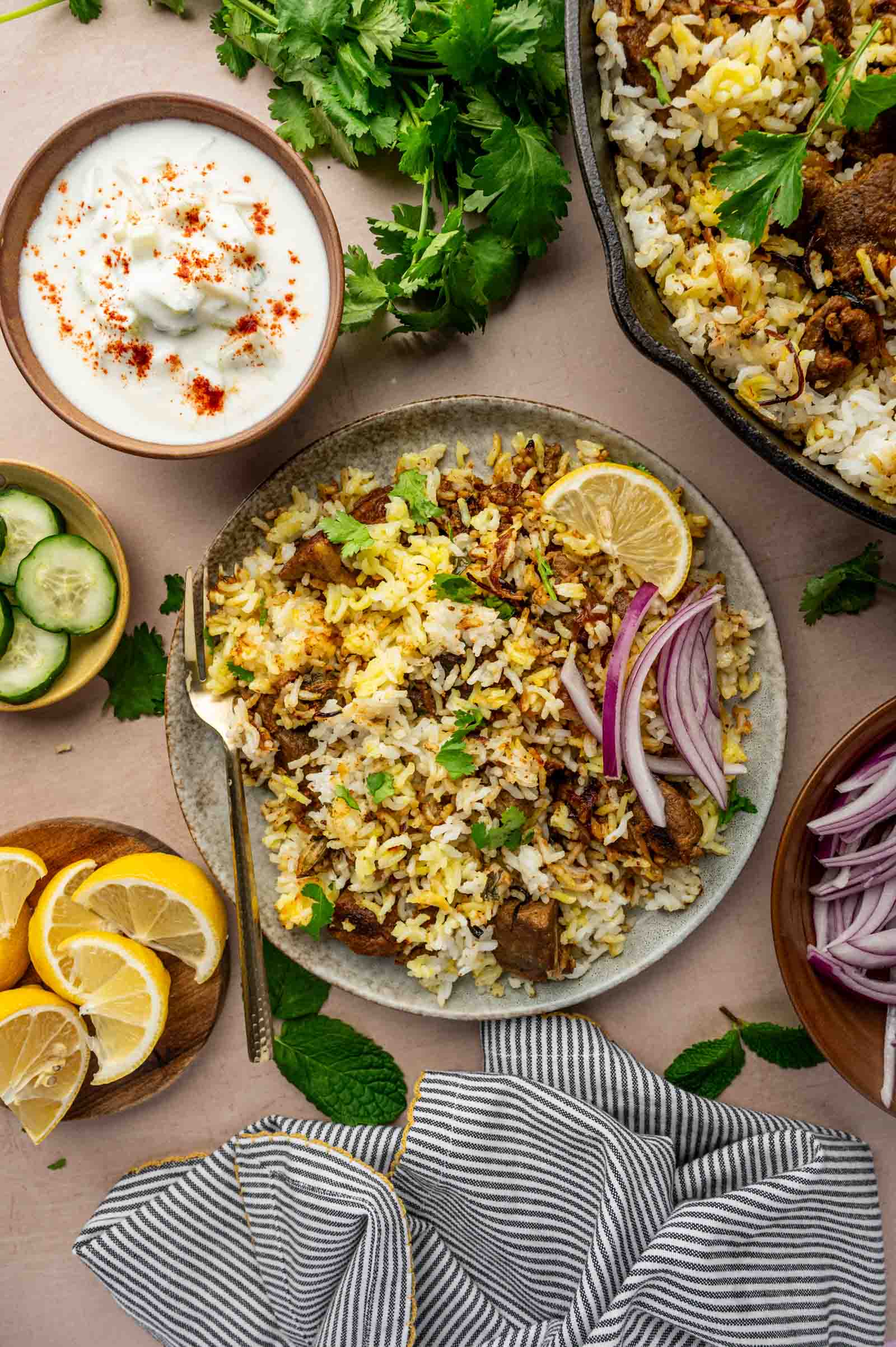 A plate of lamb biryani garnished with lemon slices, red onion, and cilantro is surrounded by a bowl of raita, cucumber slices, lemon wedges, and fresh herbs.
