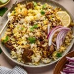 A plate of lamb biryani garnished with sliced red onions, a lemon wedge, and cilantro, with bowls of cucumber, yogurt, and extra onions on the side.