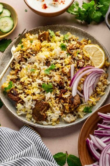 A plate of lamb biryani garnished with sliced red onions, a lemon wedge, and cilantro, with bowls of cucumber, yogurt, and extra onions on the side.