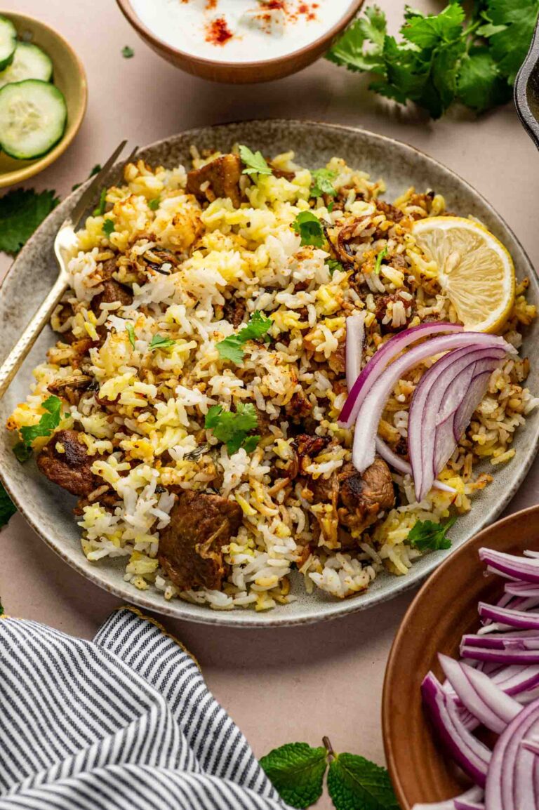A plate of lamb biryani garnished with sliced red onions, a lemon wedge, and cilantro, with bowls of cucumber, yogurt, and extra onions on the side.