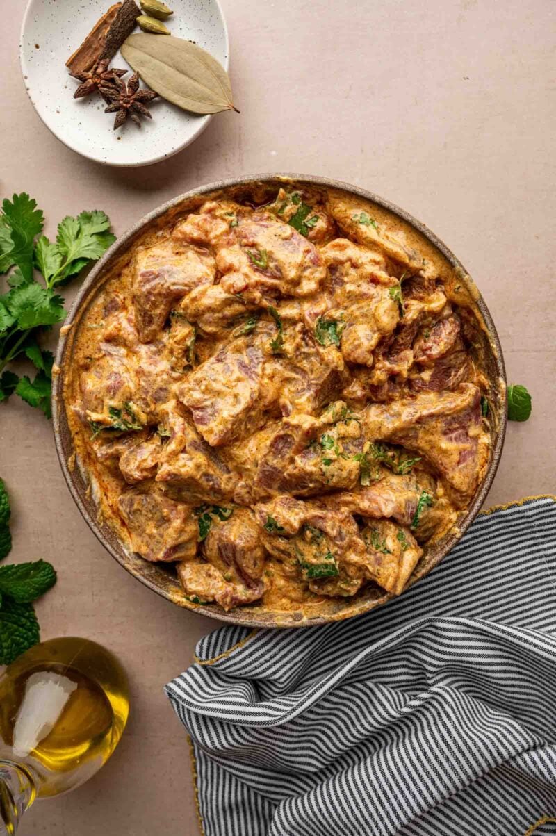 A bowl of marinated lamb with herbs and spices, placed on a beige surface next to a striped cloth, fresh herbs, whole spices, and a bottle of oil.