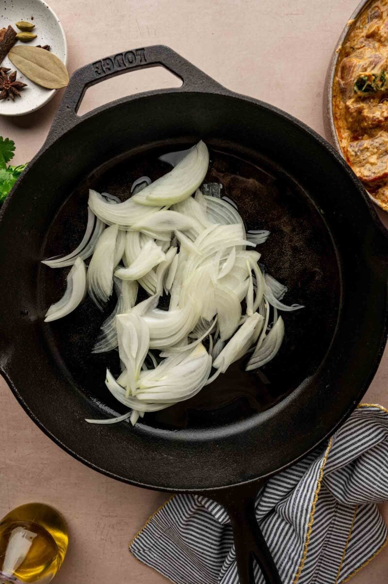 Sliced raw onions in a black cast iron skillet, ready to be cooked, with a bottle of oil, a towel, and spices nearby.