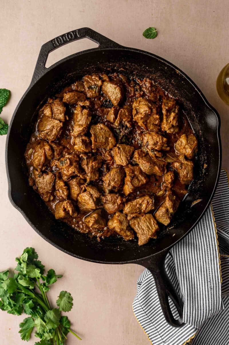 Chunks of cooked lamb in a dark sauce inside a black cast iron skillet, with cilantro and a striped cloth nearby on a light surface.