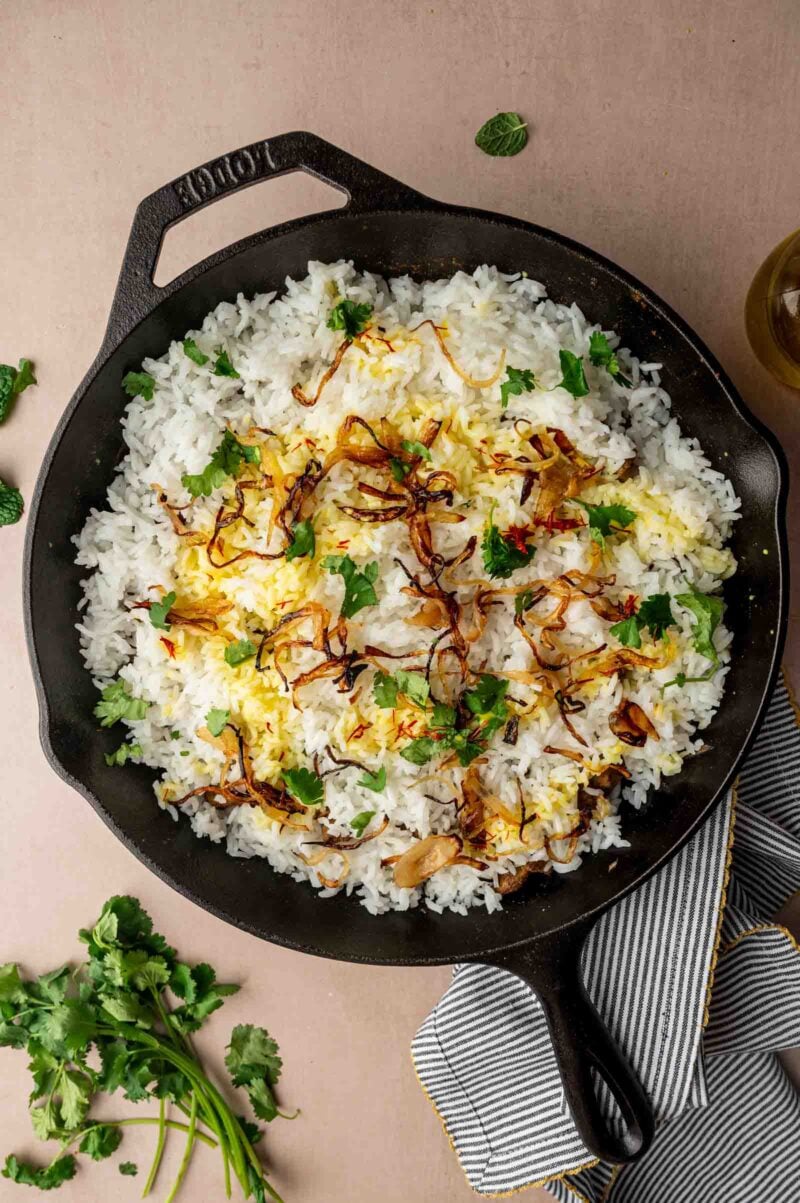 A cast iron skillet filled with rice, garnished with fried onions, fresh herbs, and a drizzle of saffron. A bunch of cilantro and a striped towel are nearby.