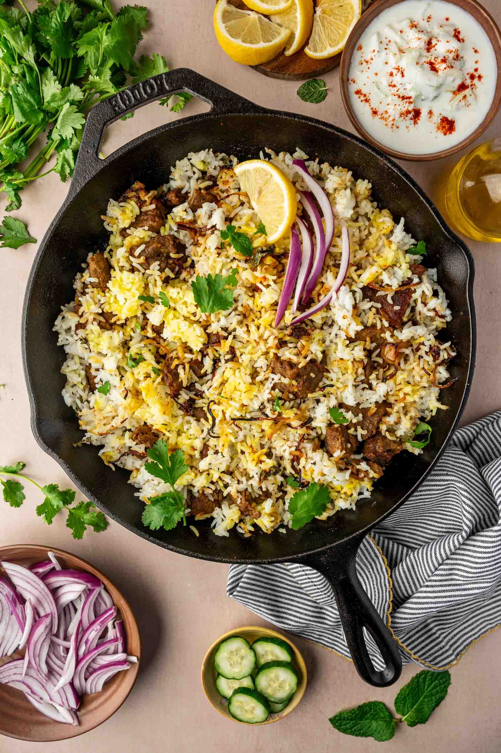 A skillet of lamb biryani garnished with lemon wedges, red onion, and cilantro, surrounded by yogurt, cucumber slices, herbs, and lemon halves.