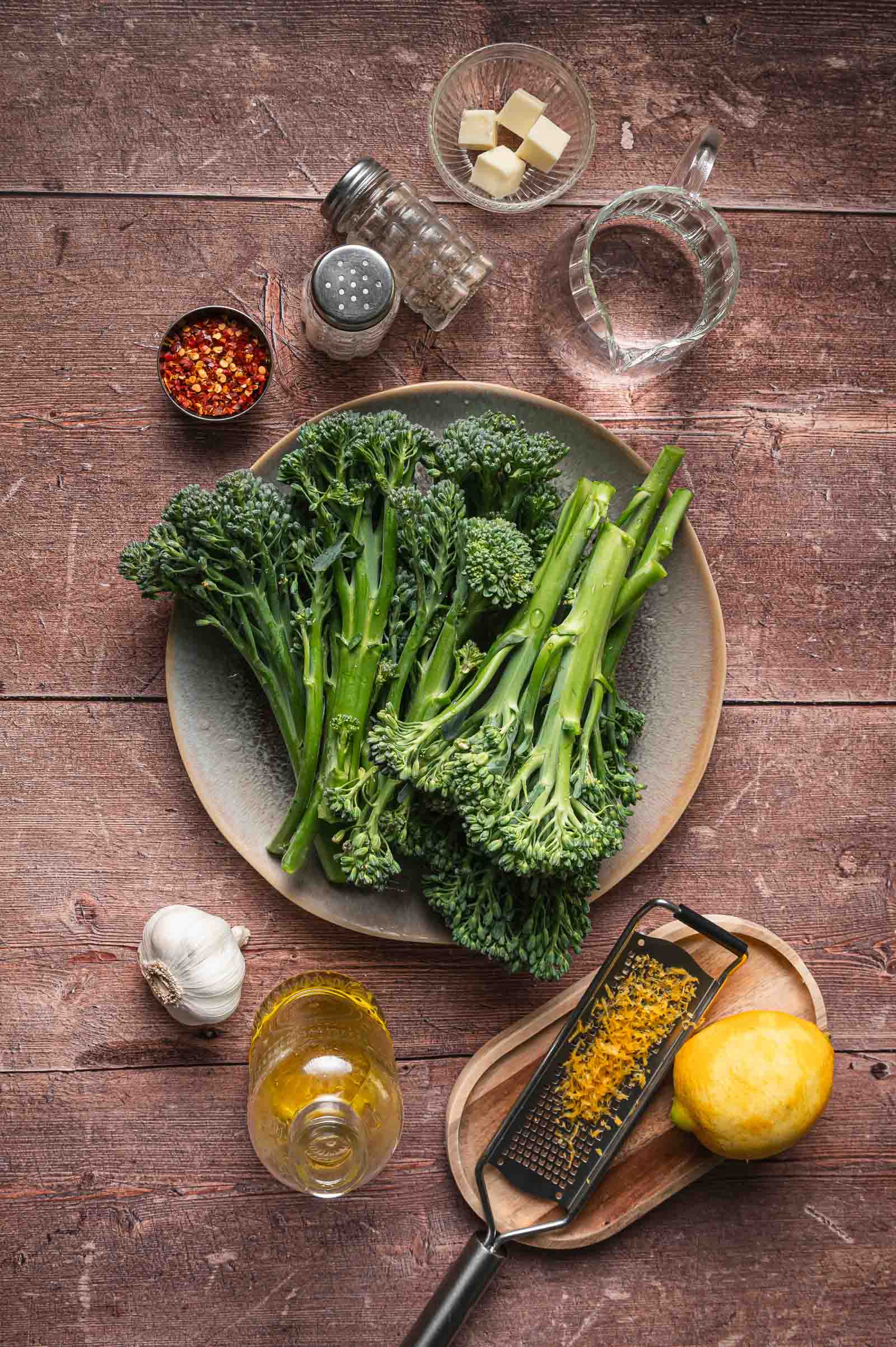 A plate of broccolini on a table surrounded by salt, red pepper flakes, butter, water, garlic, olive oil, grated lemon zest, and a lemon.