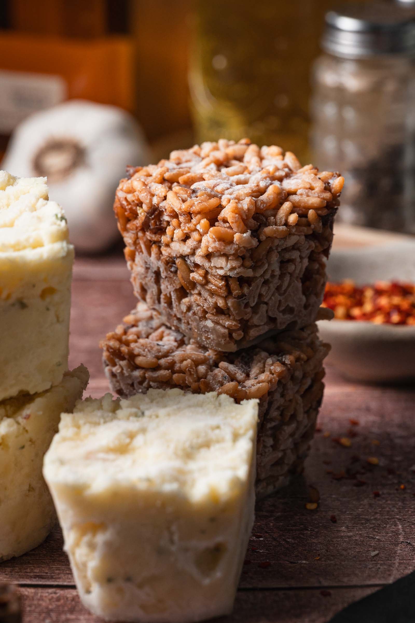Two frozen cubes of stick of butter rice and a cube of frozen boursin mashed potatoes stacked on a wooden surface, with garlic and spices blurred in the background.