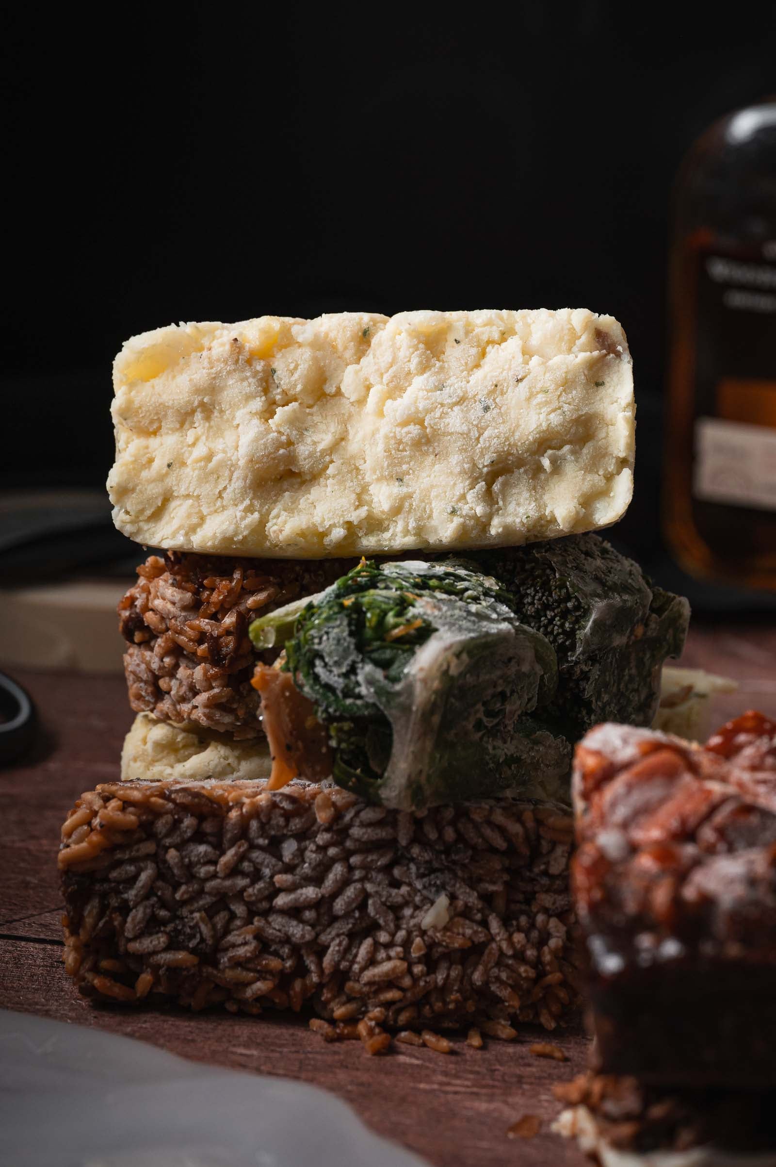 A close-up of three stacked frozen meal prep recipes - one of mashed potatoes one of lemon broccolini and one of stick of butter rice all on a wooden surface.
