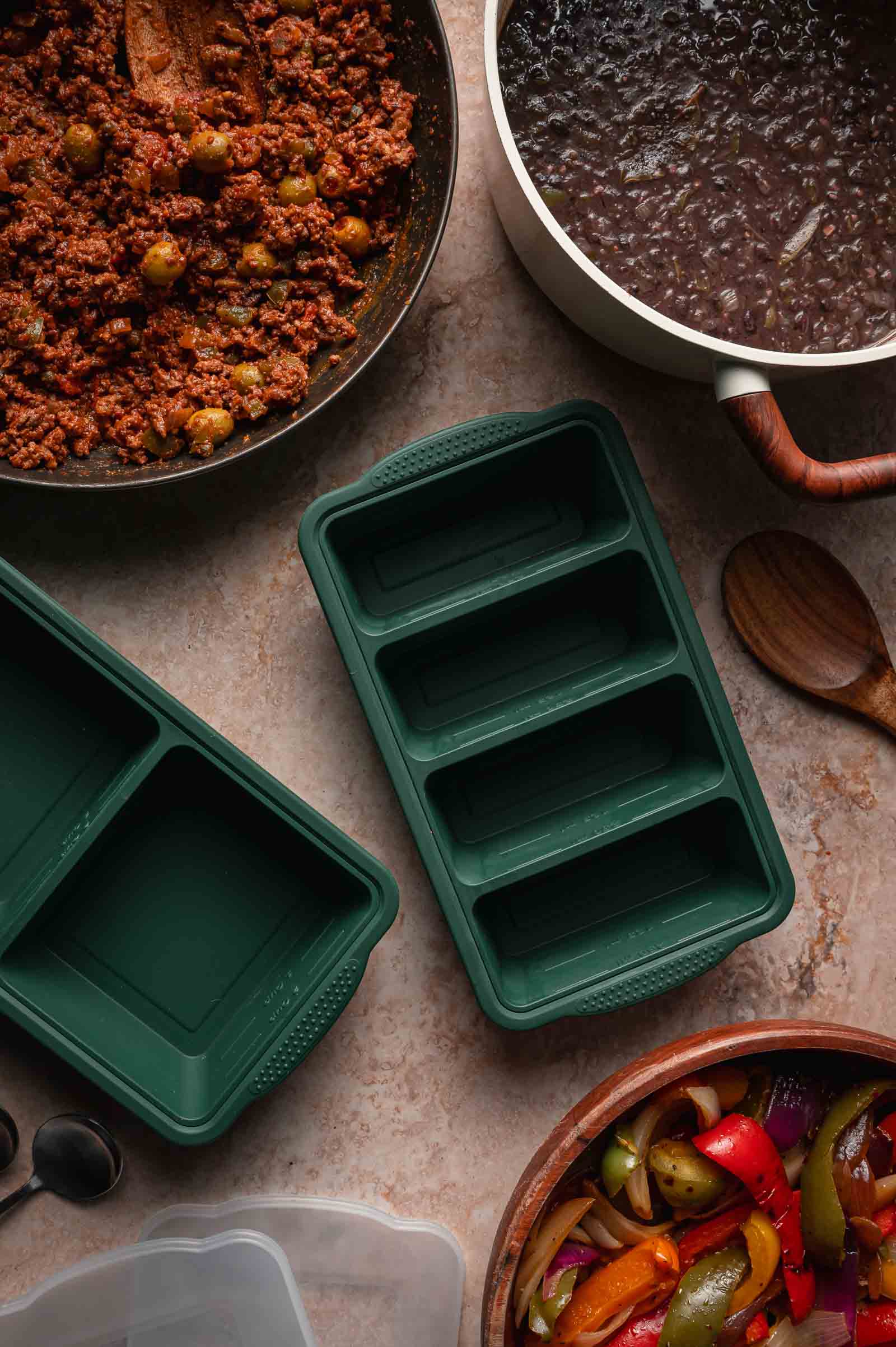 A countertop with cooked picadillo, black beans, peppers and onions, green silicone meal prep containers, and a wooden spoon.