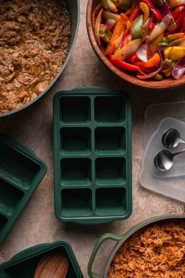 A green silicone freezer tray surrounded by cooked mississippi chicken, peppers and onions, and spanish rice on a beige countertop, with two metal spoons nearby.