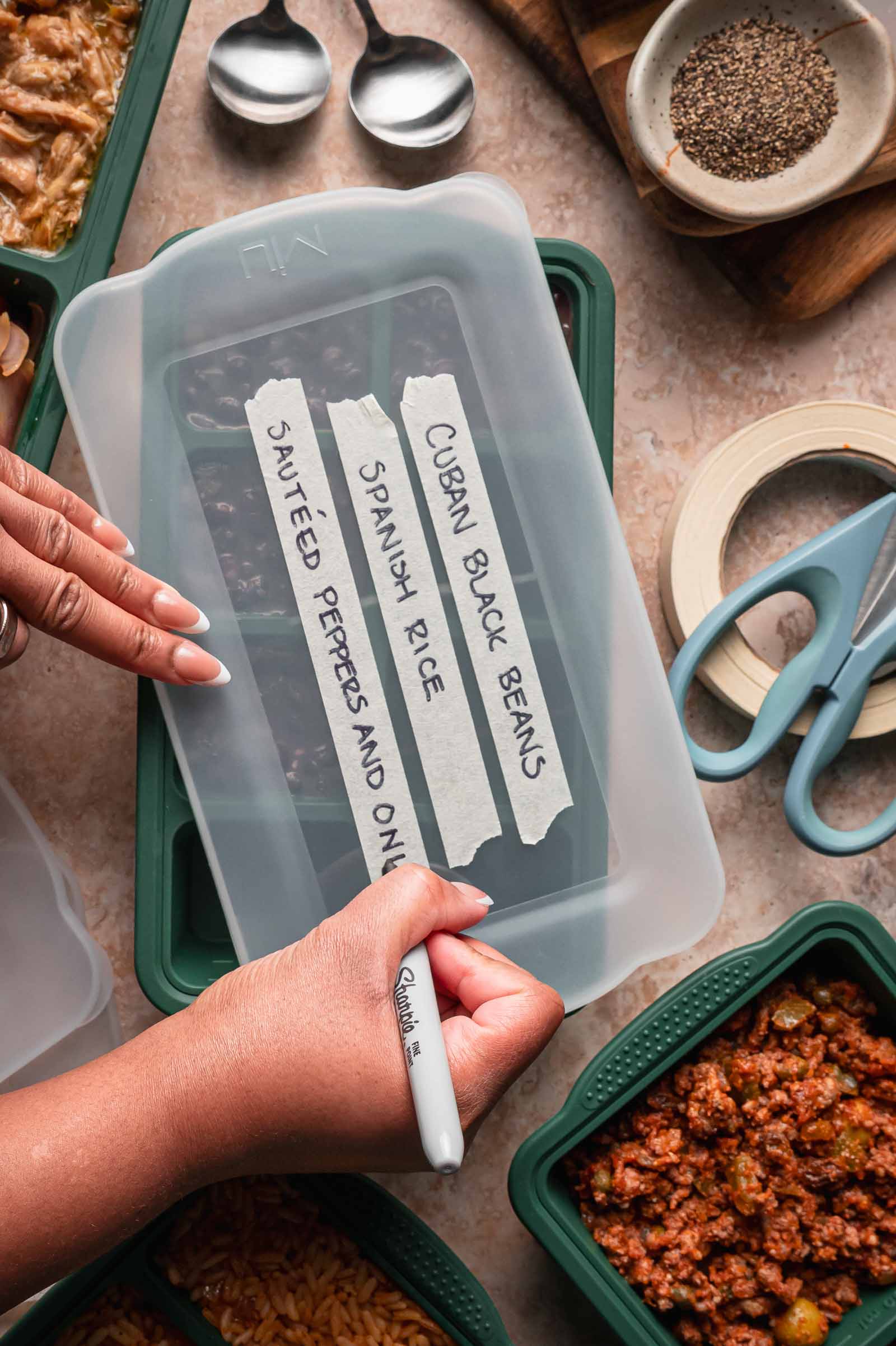 A person labels a silicone meal prep food container lid with a marker. The container holds Spanish rice, Cuban black beans, and sautéed peppers and onions. Prepared food and kitchen tools surround the scene.