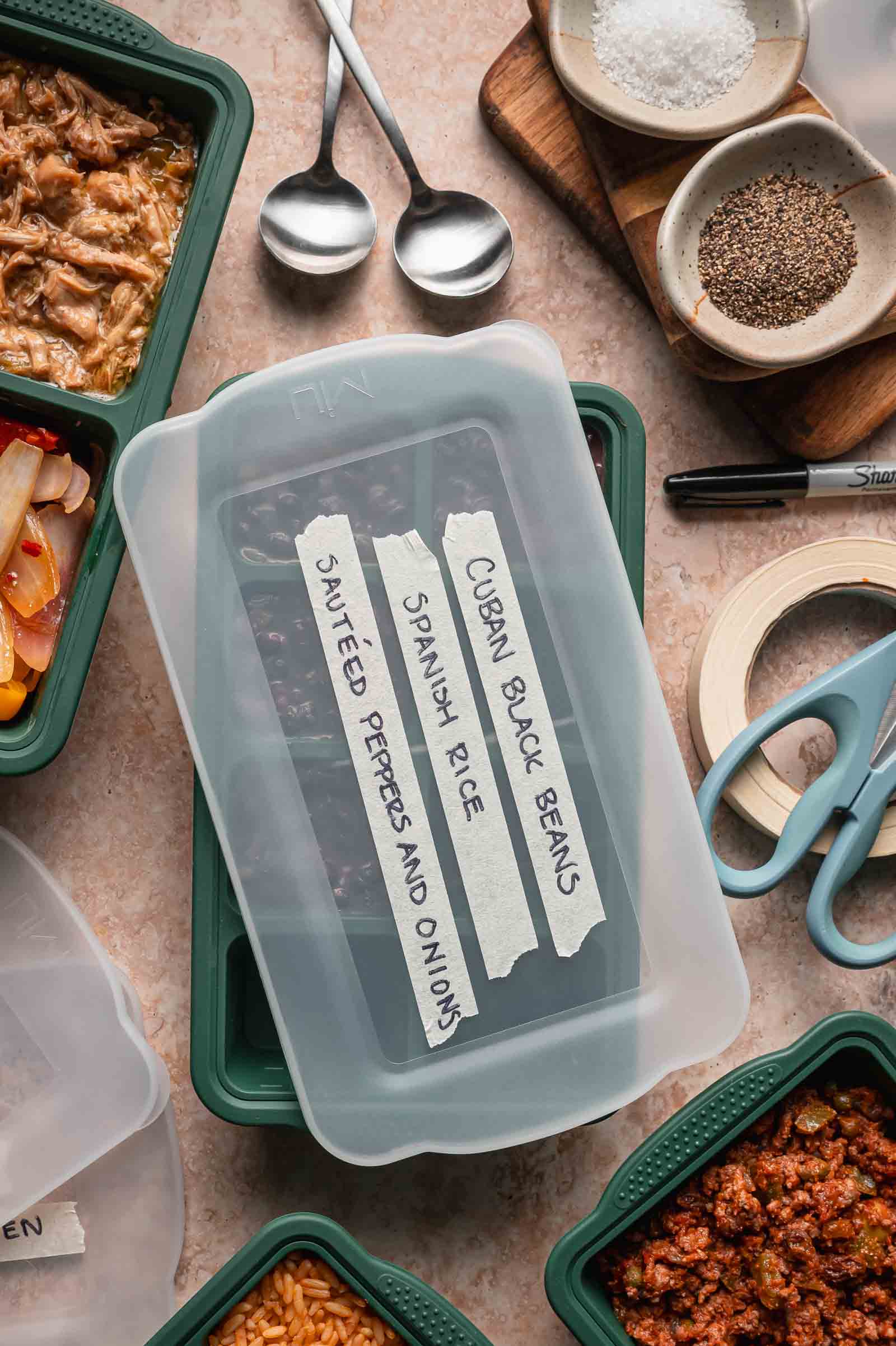 Rectangular food container with three labeled sections: “Sautéed Peppers and Onions,” “Spanish Rice,” and “Cuban Black Beans,” surrounded by meal prep items like spoons, spices, and scissors.