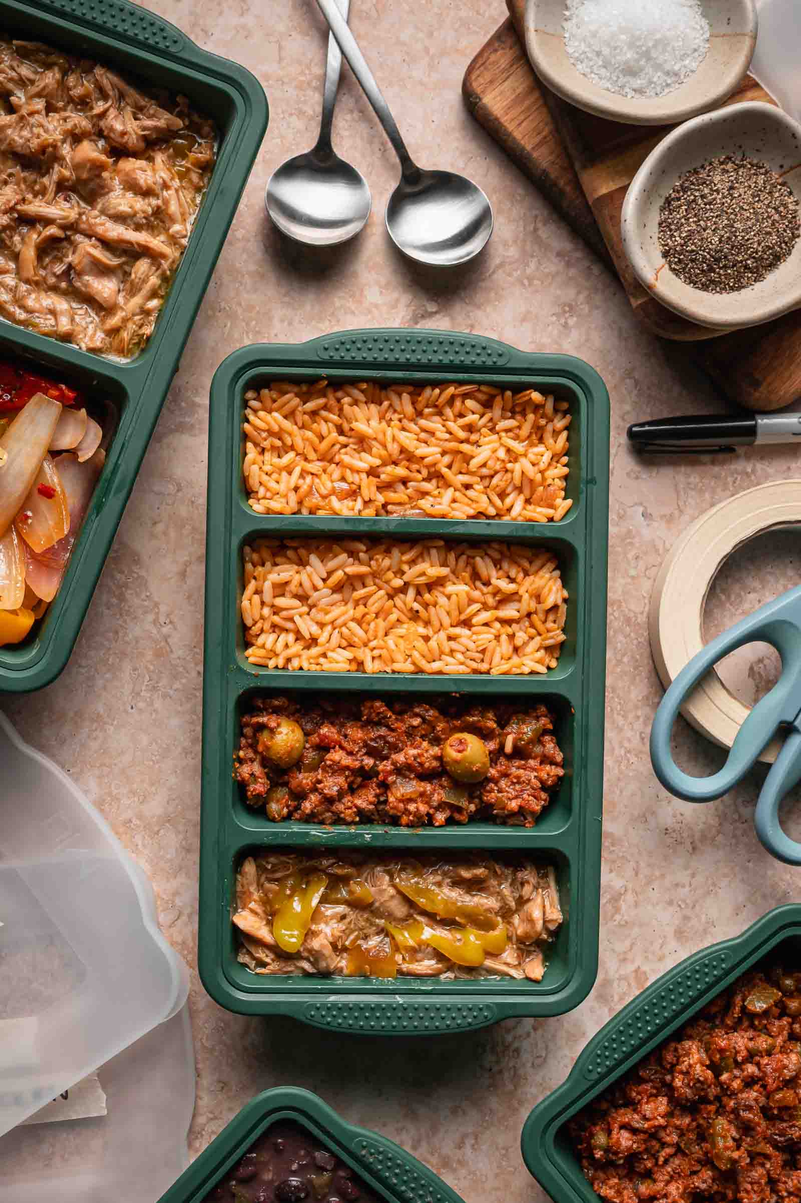 A green meal prep container with three sections holds Spanish rice, picadillo, and mississippi chicken. Spoons, spices, and kitchen tools are nearby on the countertop.