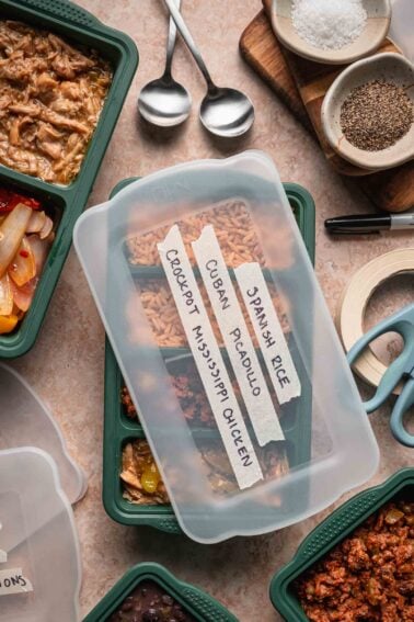 A labeled meal prep container with three compartments of food is surrounded by other containers, utensils, seasonings, and kitchen tools on a countertop.