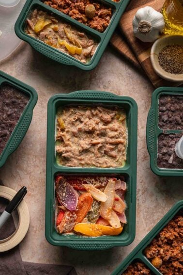 A green meal container holds shredded Mississippi chicken and peppers and onions; other containers with various foods surround it on a beige countertop.