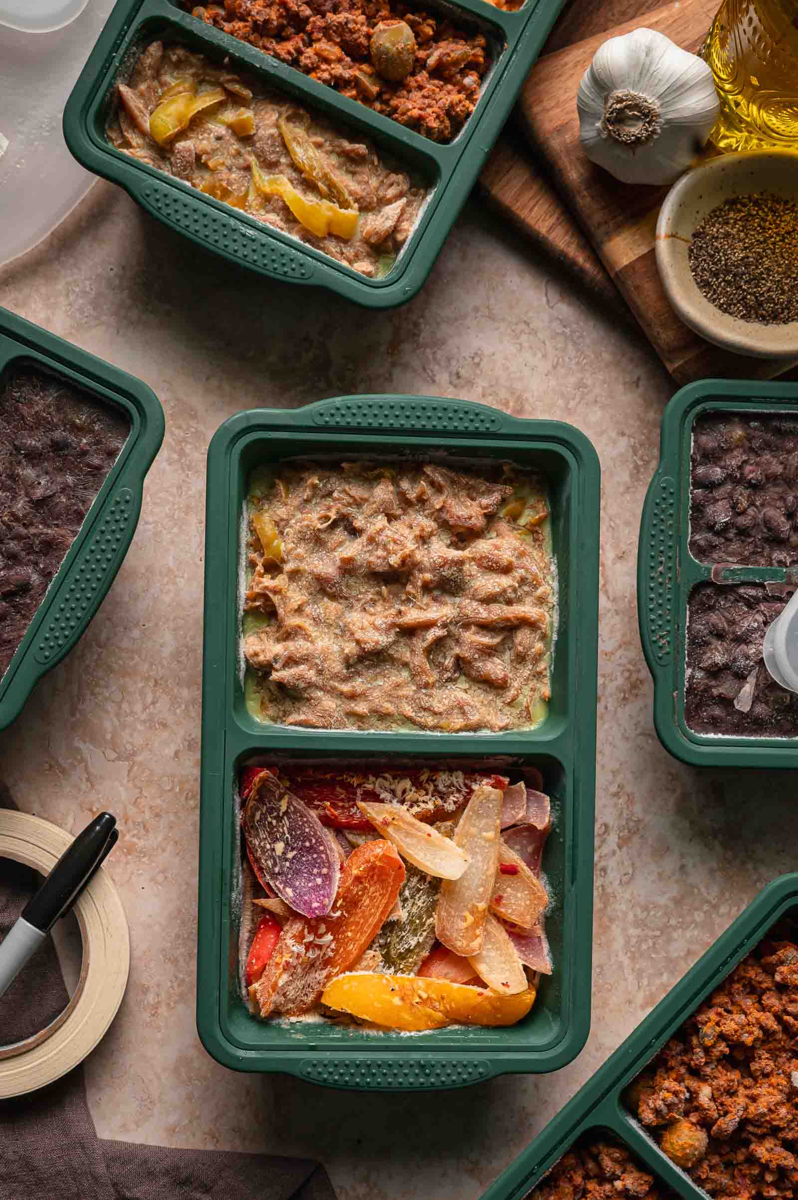 A green meal container holds shredded Mississippi chicken and peppers and onions; other containers with various foods surround it on a beige countertop.