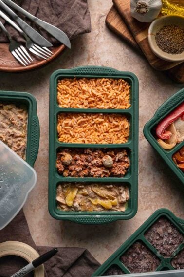 A green meal prep tray with frozen compartments containing Spanish rice, picadillo, and a Mississippi chicken, surrounded by kitchen utensils and ingredients.