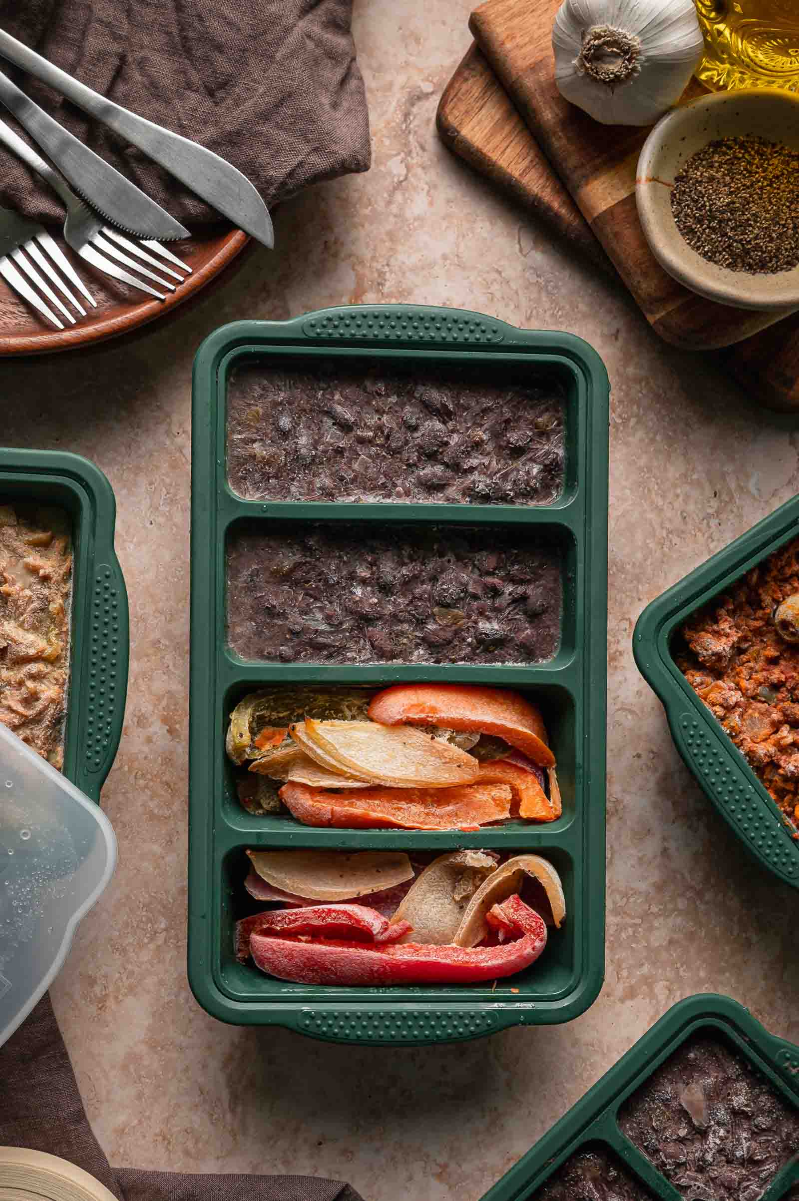 A divided green silicone meal prep container with frozen Cuban black beans and peppers and onions on a countertop surrounded by utensils, spices, and other meal containers.