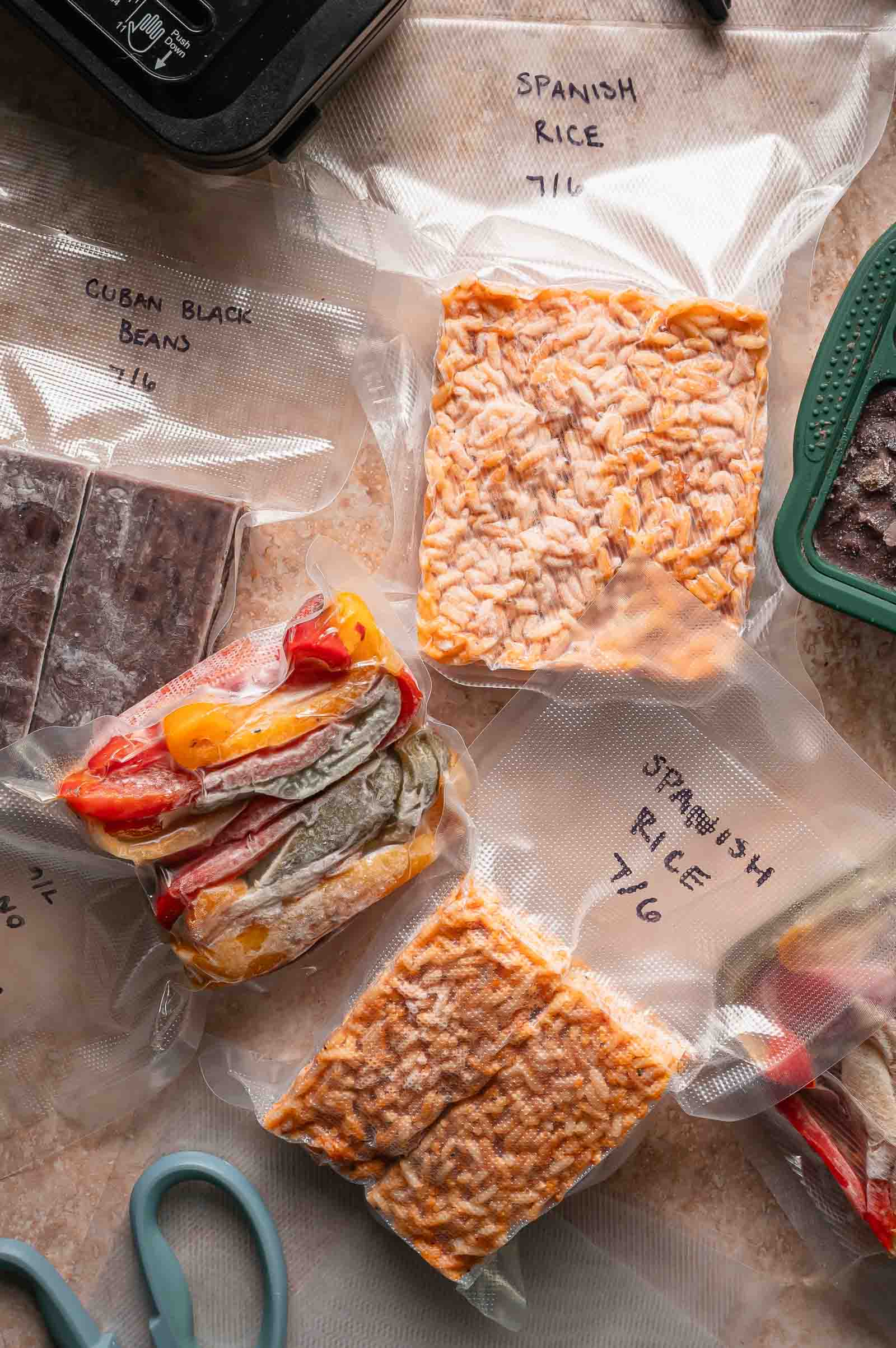 Vacuum-sealed bags of Spanish rice, Cuban black beans, and peppers and onions are labeled and arranged on a countertop with scissors and a portion of a green container visible.