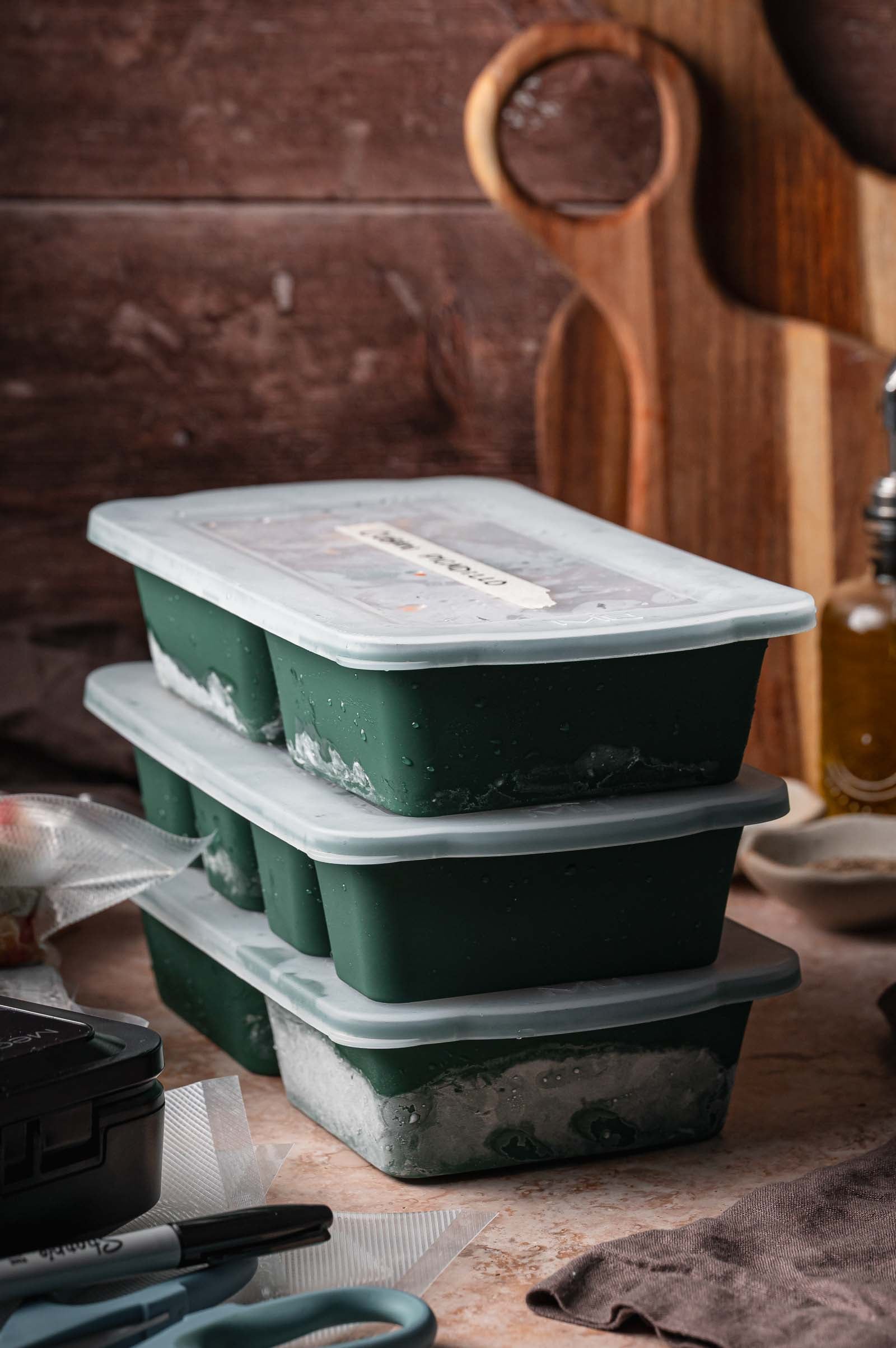 Three frozen green silicon meal prep containers are stacked on a kitchen counter with cutting boards and utensils in the background.