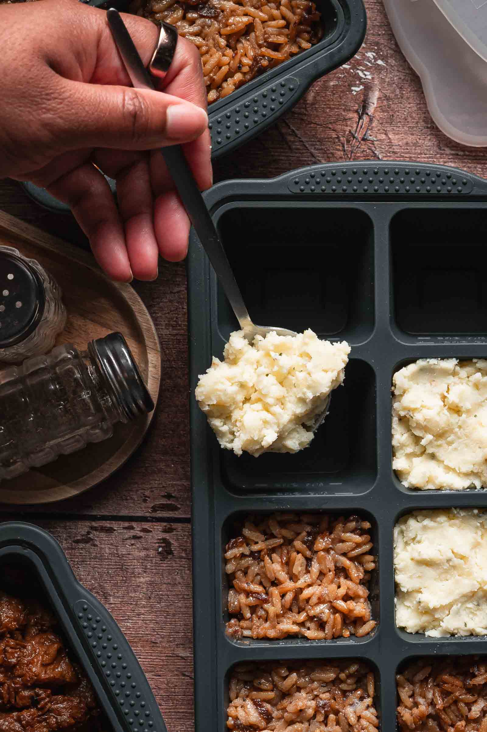 A hand uses a spoon to add mashed potatoes to a sectioned silicon mold for meal prepping, with rice and other prepared food in adjacent sections.