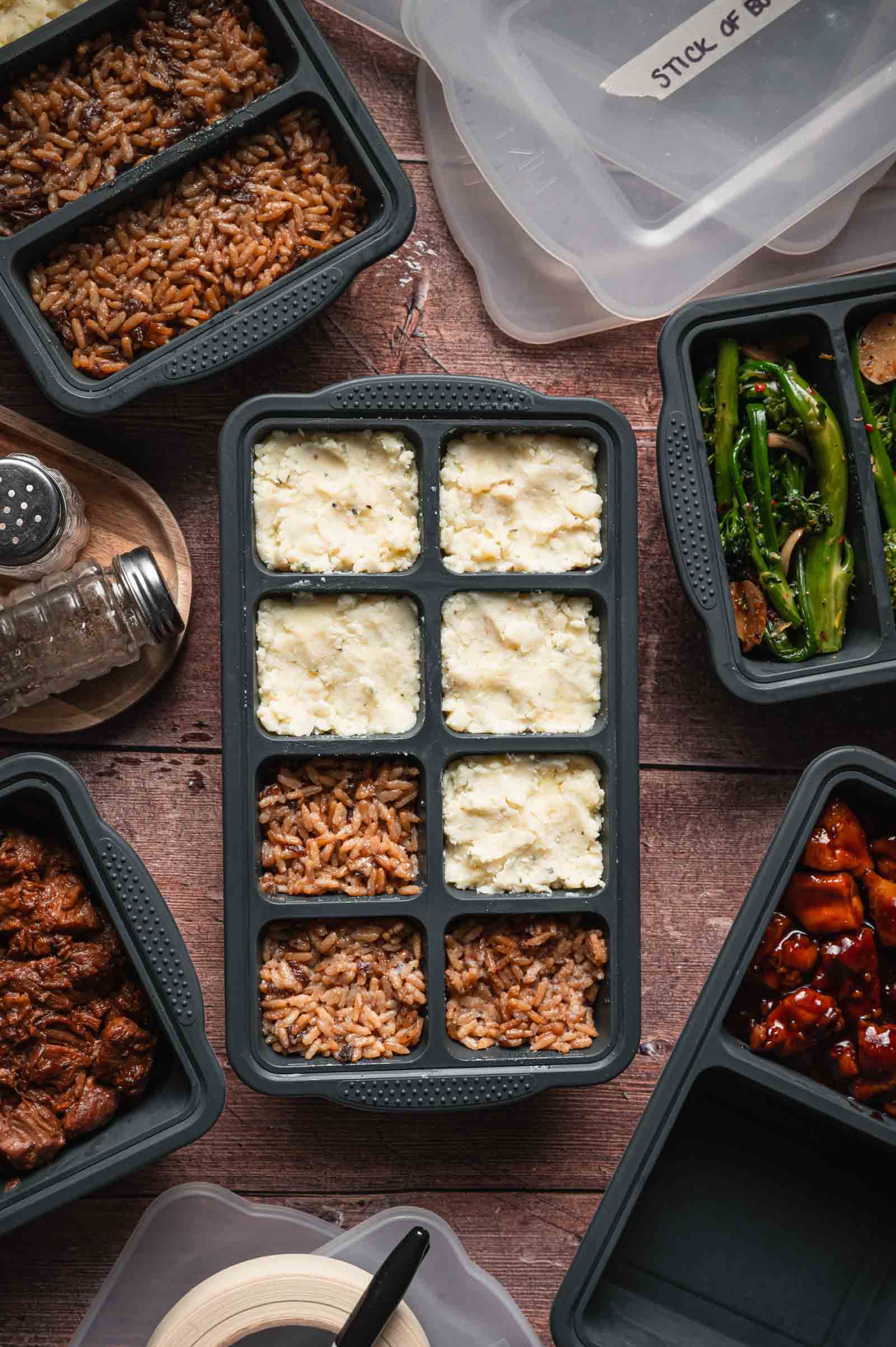 A meal prep container filled with mashed potatoes and stick of butter rice, surrounded by additional containers of cooked vegetables and meat in similar containers on a wooden surface.