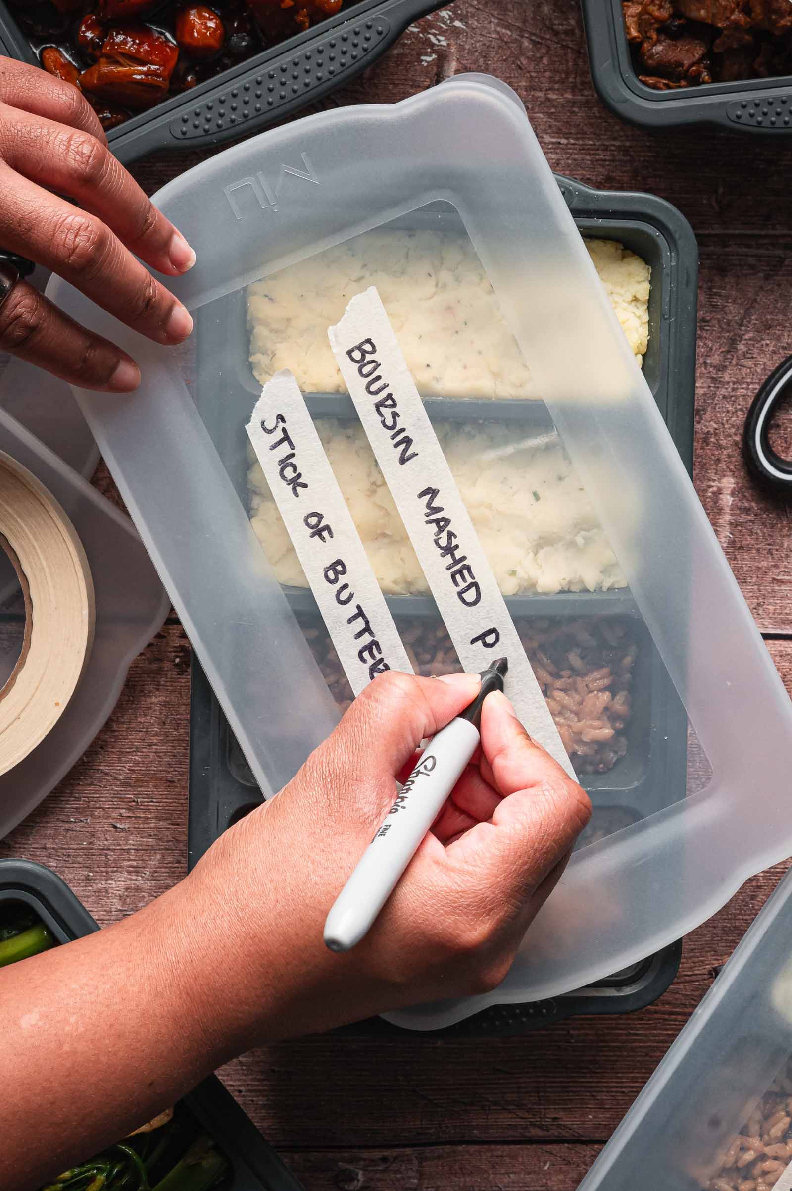 A person labels a plastic food container with a marker, writing “Boursin Mashed” and “Stick of Butter.” Other meal prep containers are on the table nearby.