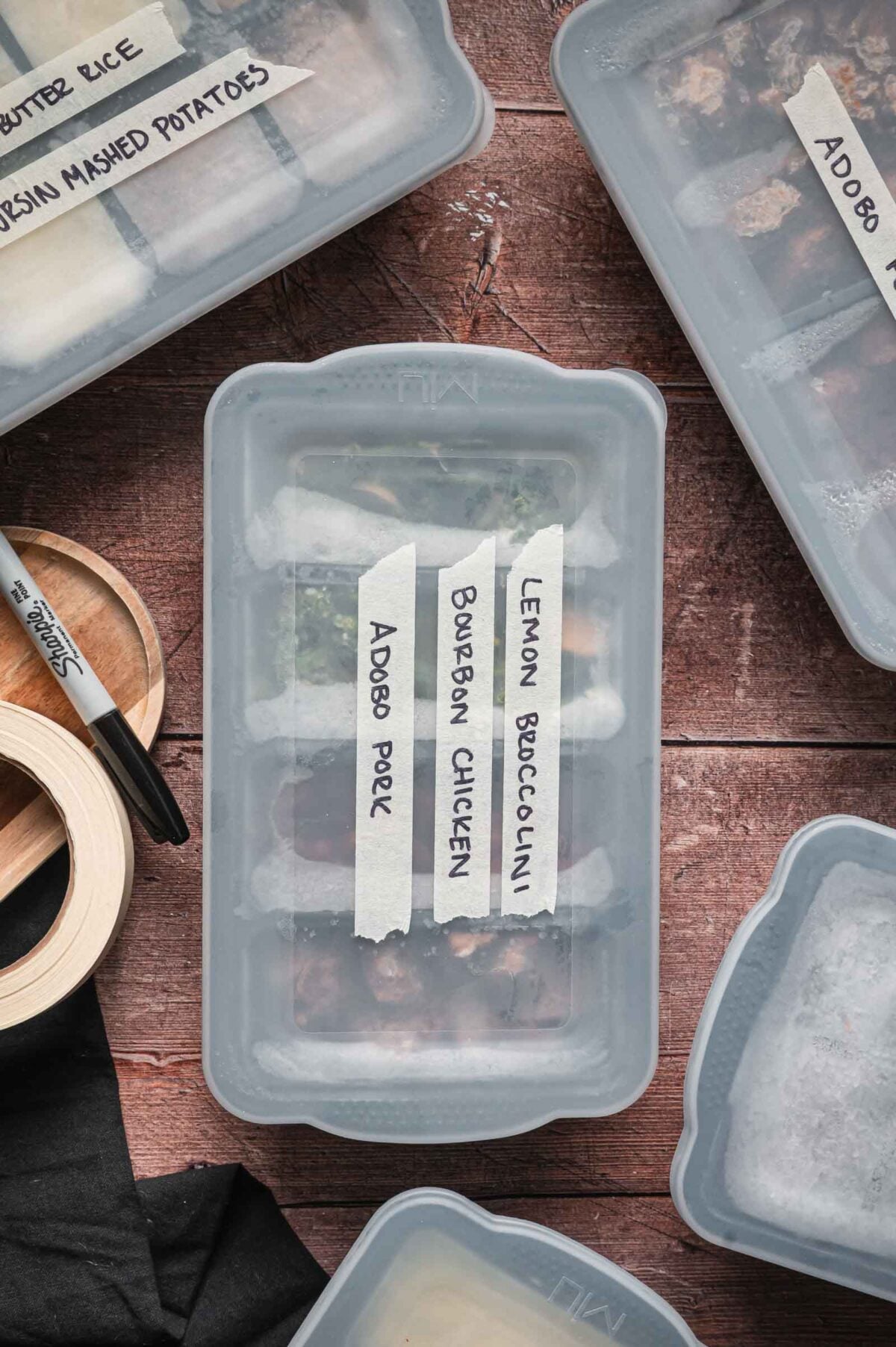 Several labeled meal prep containers with handwritten tape labels, including Adobo Pork, Bourbon Chicken, and Lemon Broccolini, are arranged on a wooden surface.