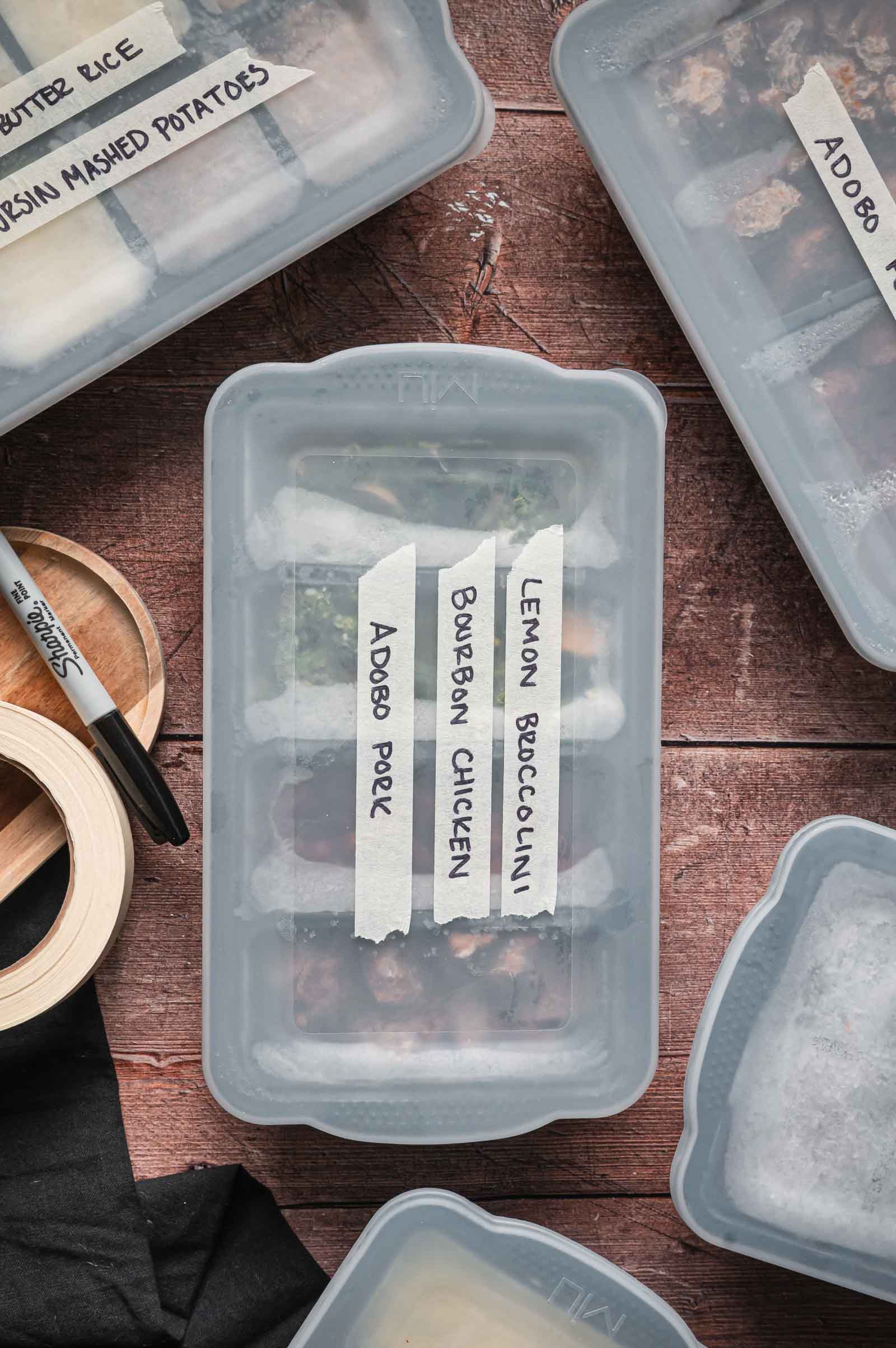 Several labeled meal prep containers with handwritten tape labels, including “Adobo Pork,” “Bourbon Chicken,” and “Lemon Broccolini,” are arranged on a wooden surface.