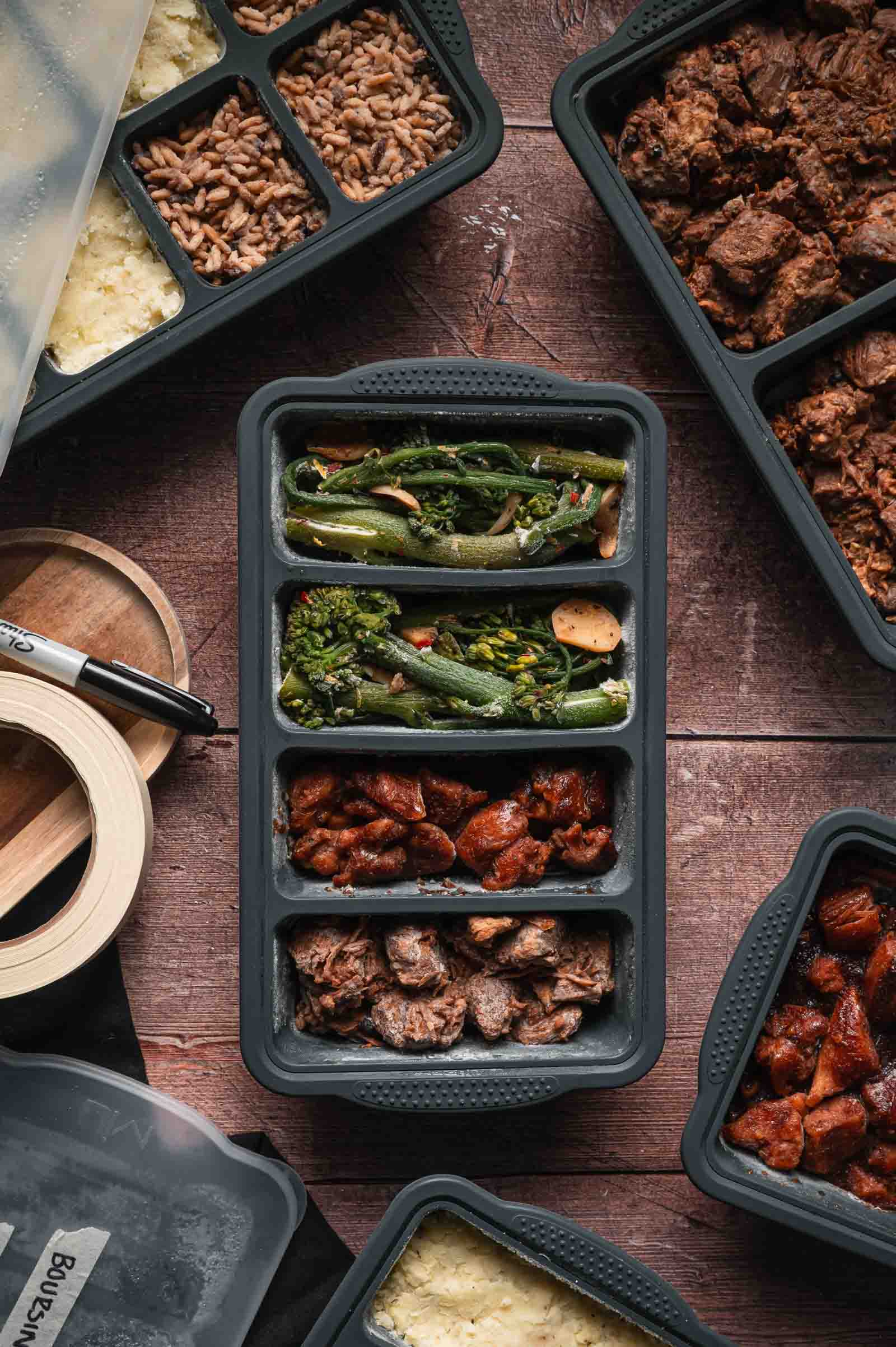 A meal prep container with sections holding frozen cookied vegetables, bourbon chicken and adobo pork sits on a wooden surface, surrounded by other similar meal containers.