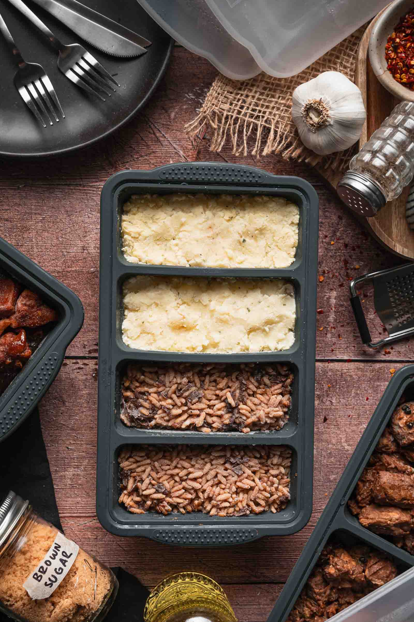A divided meal prep container holding mashed potatoes, and stick of butter rice, surrounded by spices, utensils, and meal prep items on a wooden surface.