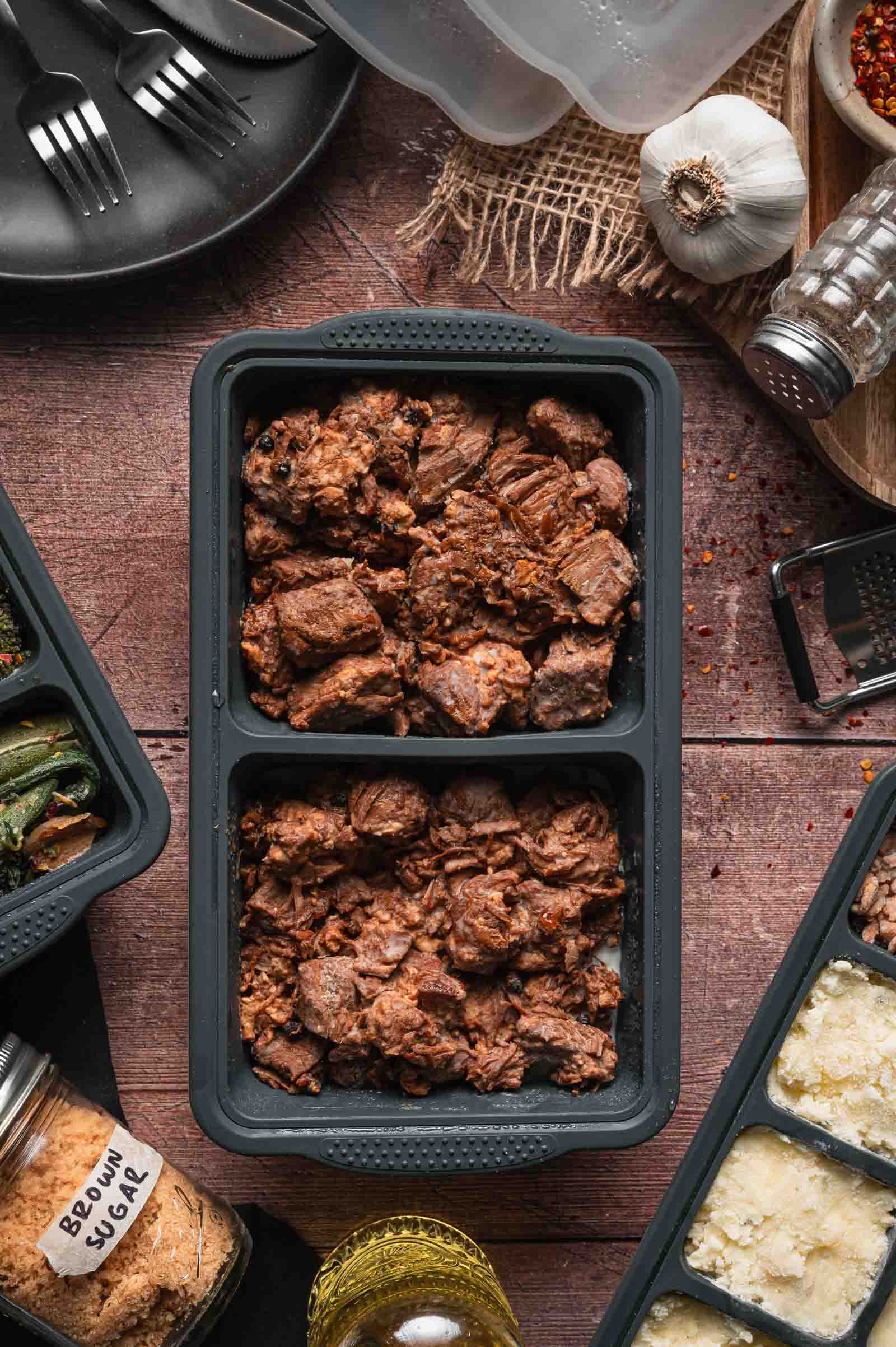 A black meal prep container with cooked adobo pork, surrounded by spices, condiments, garlic, and containers with other food items on a wooden surface.