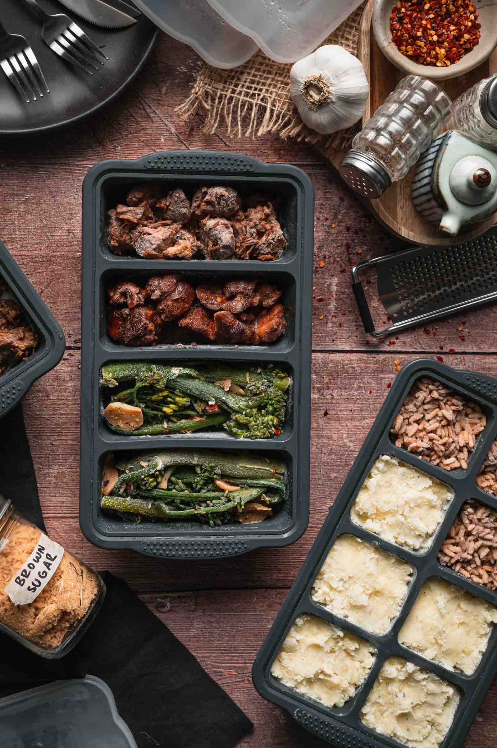 Meal prep containers with sections of frozen cooked bourbon chicken, pork adobo, broccolini, mashed potatoes and stick of butter rice, sits on a wooden table surrounded by kitchen items and seasonings.