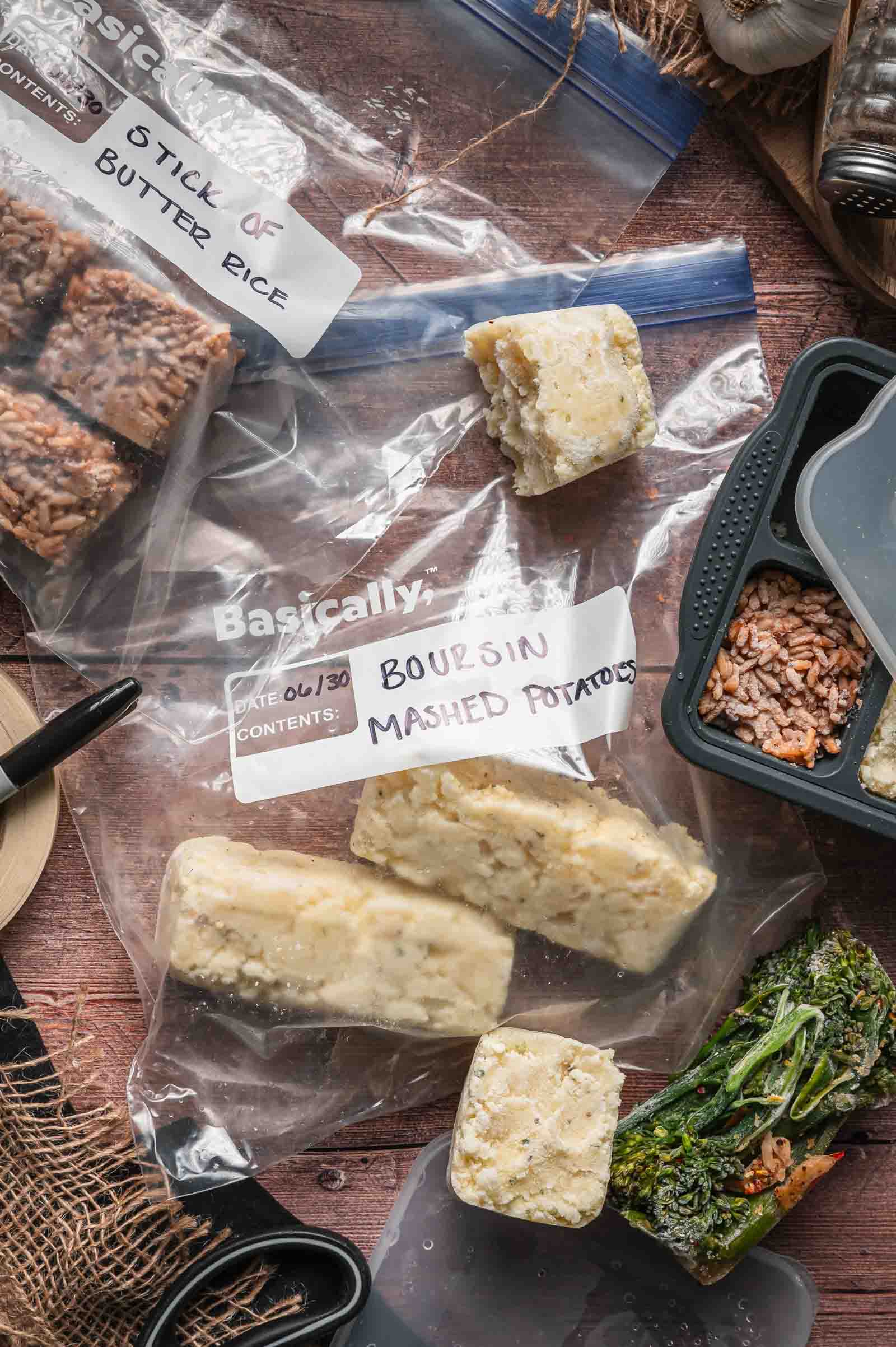 Labeled freezer bags with frozen cubes of stick of butter rice and Boursin mashed potatoes, alongside a meal tray with rice and broccoli, on a wooden surface.
