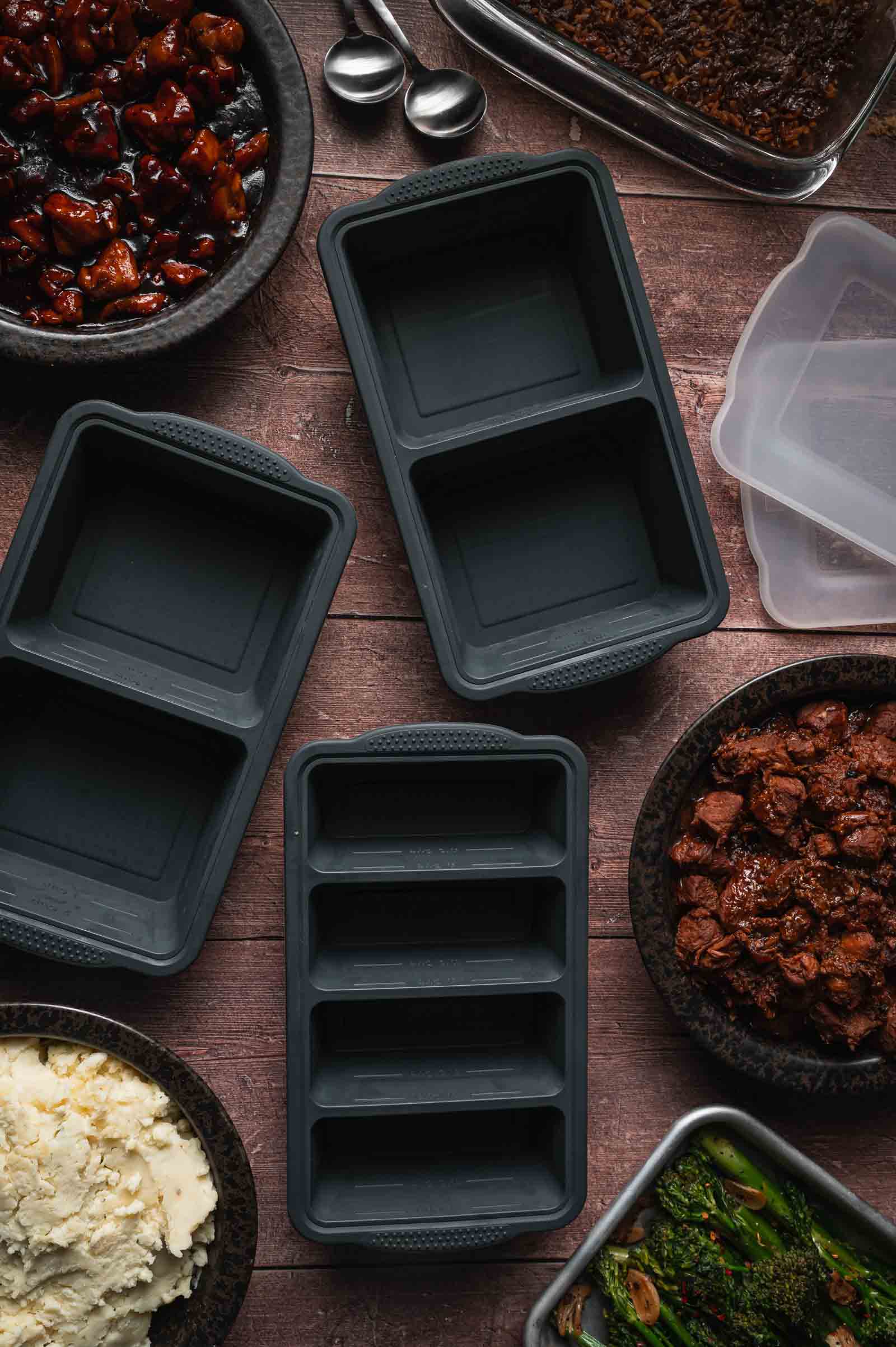 Three black meal prep containers sit on a wooden table surrounded by bowls of cooked food, including mashed potatoes, vegetables, and meat dishes.