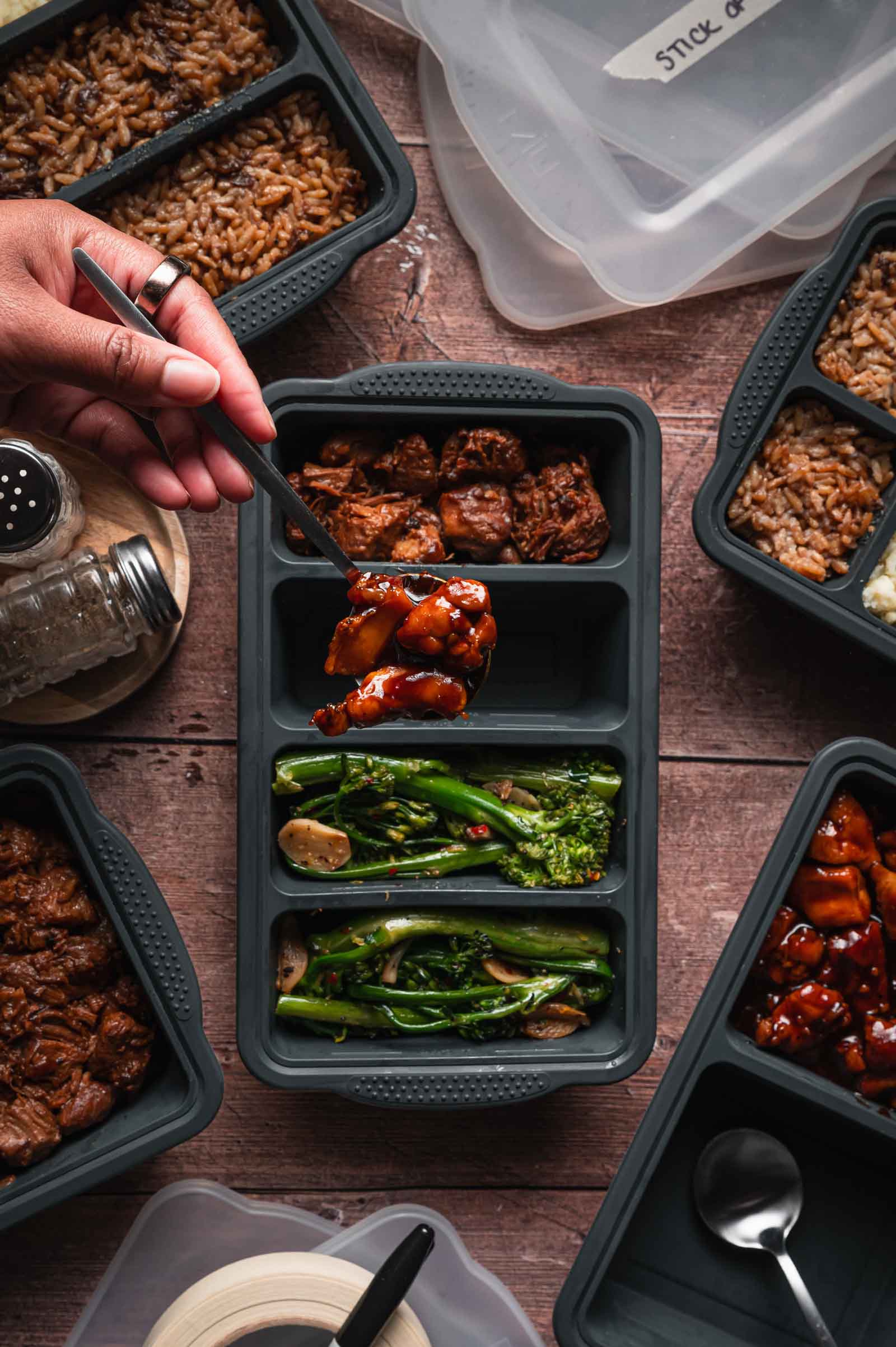 A hand holds a fork with bourbon chicken over a meal prep container filled with adobo pork, broccoli, and rice; other similar containers are arranged on a wooden table.
