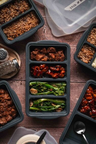 Overhead view of multiple meal prep containers with sections of rice, cooked meat, and vegetables on a wooden surface, surrounded by lids and utensils.