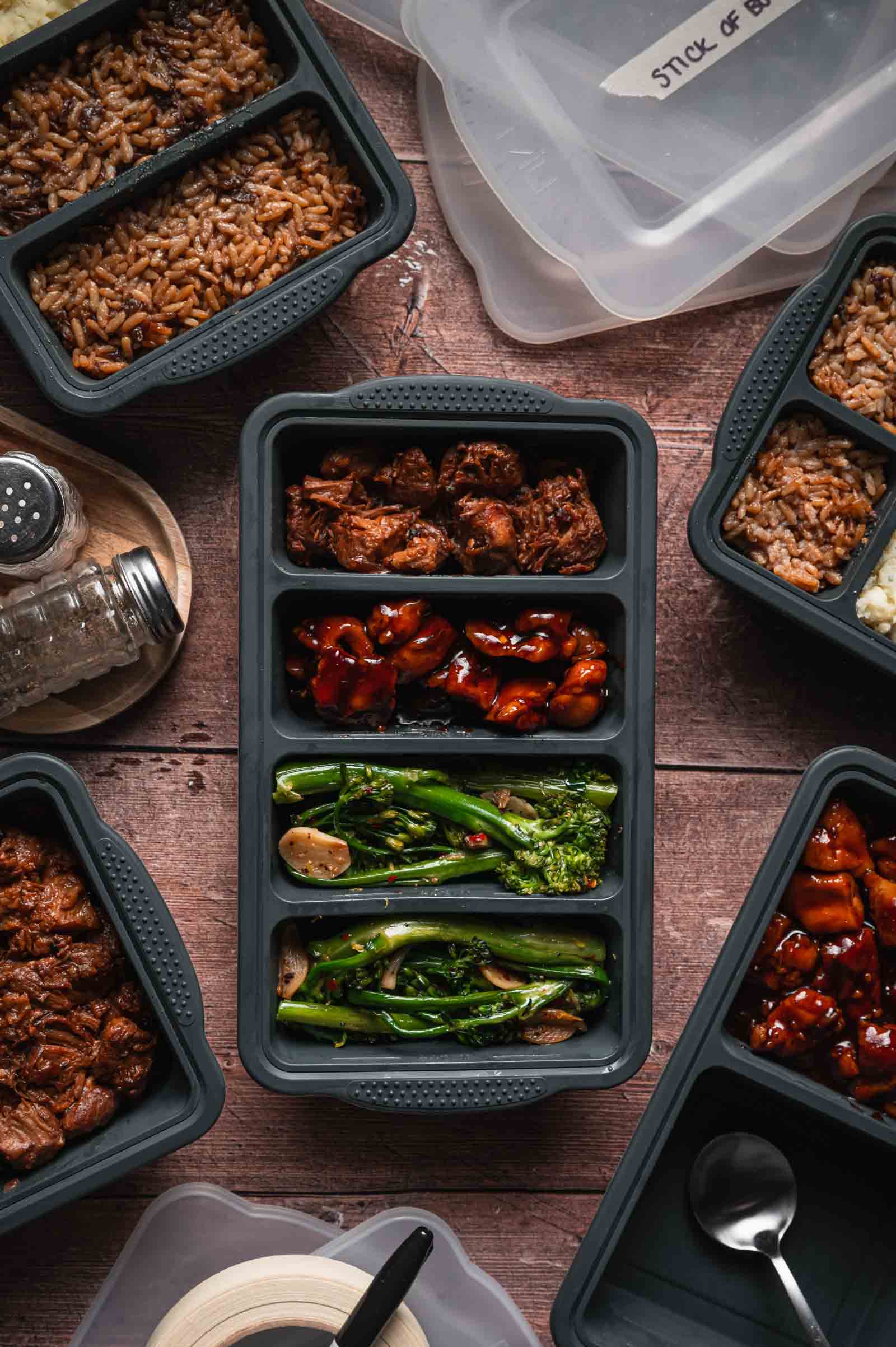 Overhead view of multiple meal prep containers with sections of rice, cooked meat, and vegetables on a wooden surface, surrounded by lids and utensils.