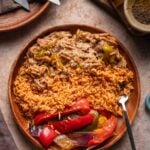 A wooden plate with Spanish rice, shredded Mississippi chicken with peppers, and peppers and onions, placed on a stone surface with utensils, garlic, and spices nearby.
