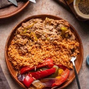 A wooden plate with Spanish rice, shredded Mississippi chicken with peppers, and peppers and onions, placed on a stone surface with utensils, garlic, and spices nearby.