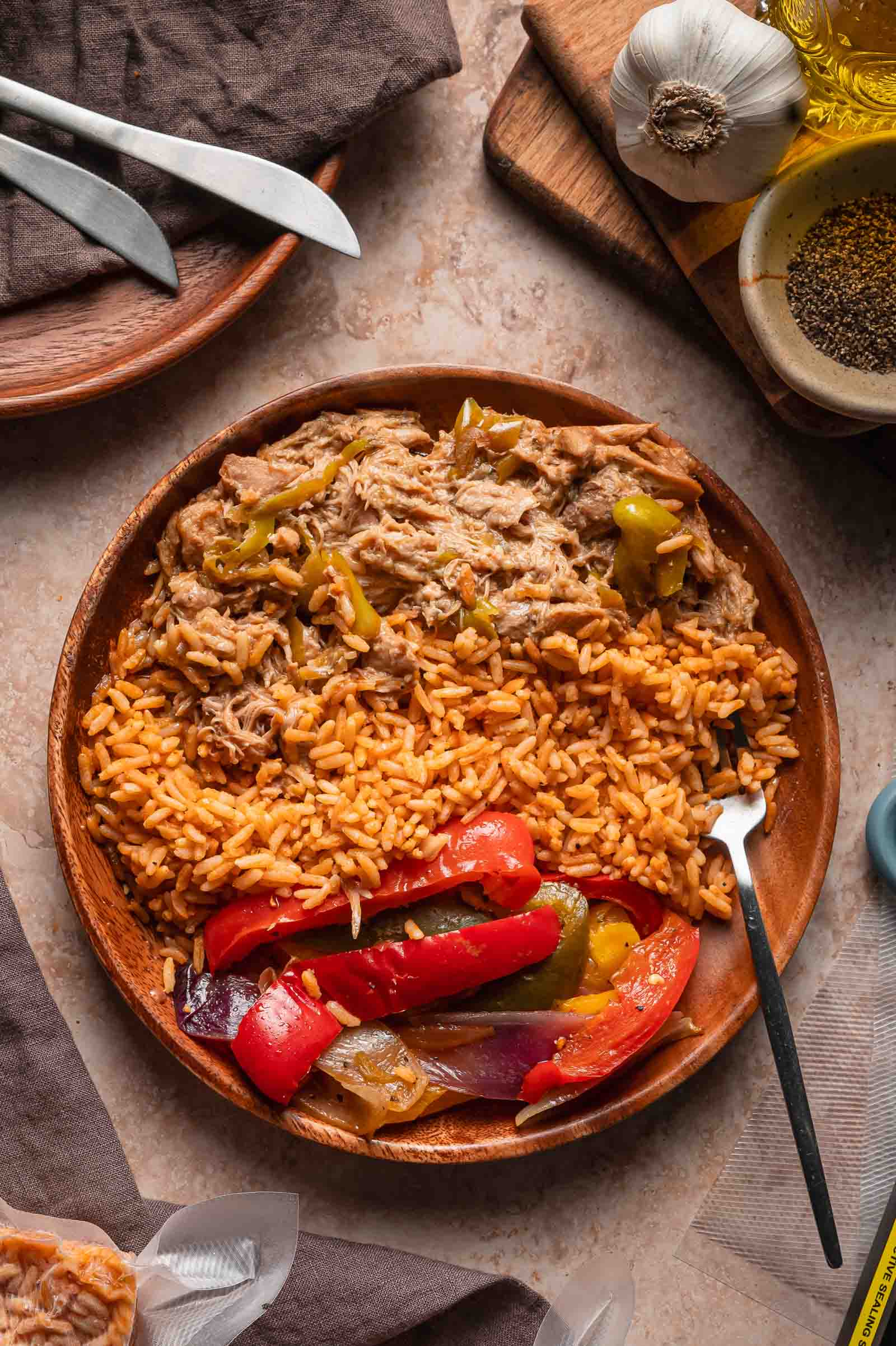 A wooden plate with Spanish rice, shredded Mississippi with peppers, and peppers and onions, placed on a stone surface with utensils, garlic, and spices nearby.